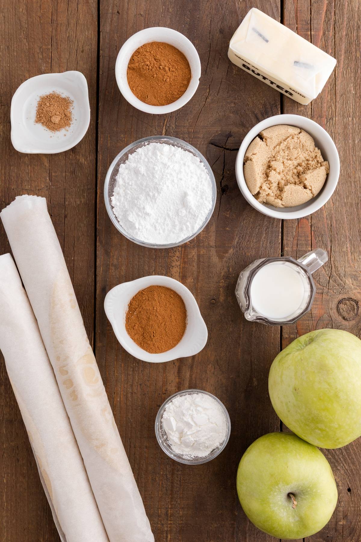 Ingredients for baking, including apples, pie crusts, spices, brown sugar, powdered sugar, butter, and milk on a table.