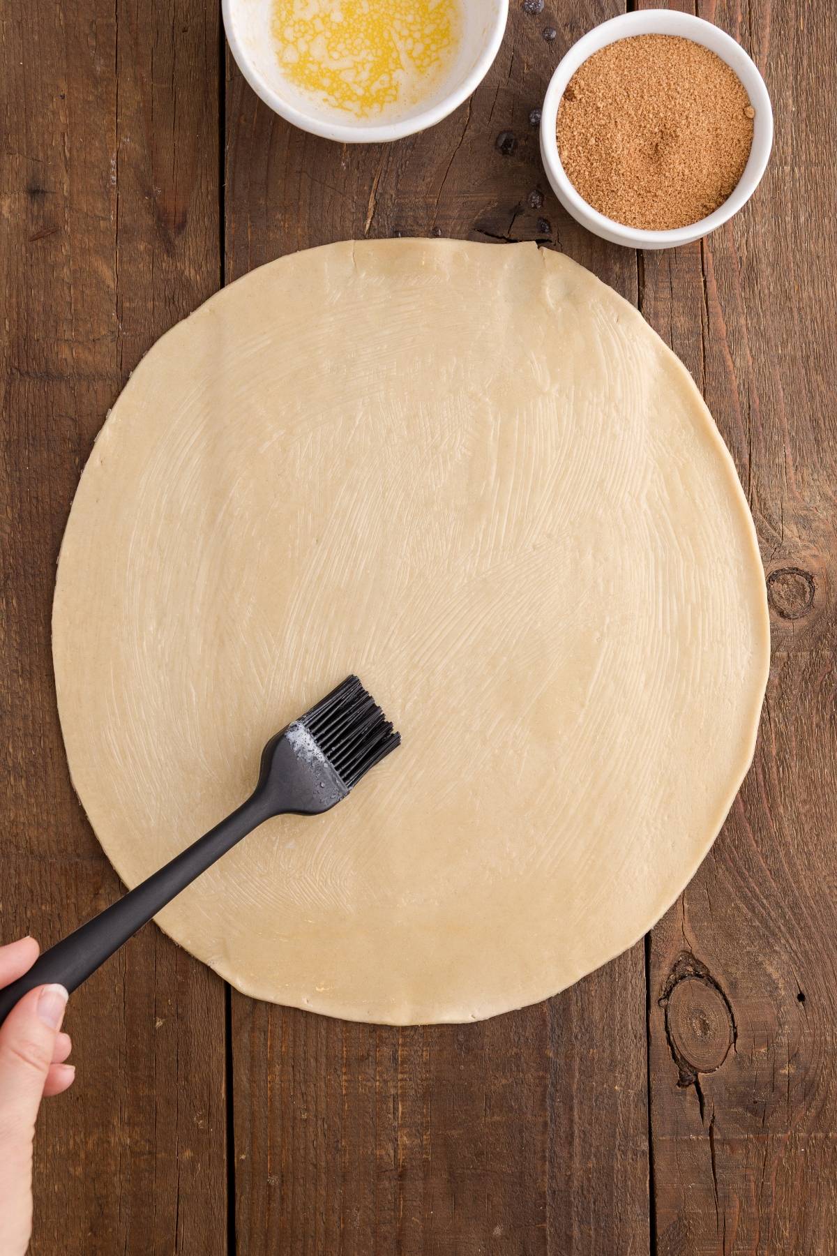 A hand brushes melted butter on rolled dough, with bowls of butter and cinnamon sugar nearby.
