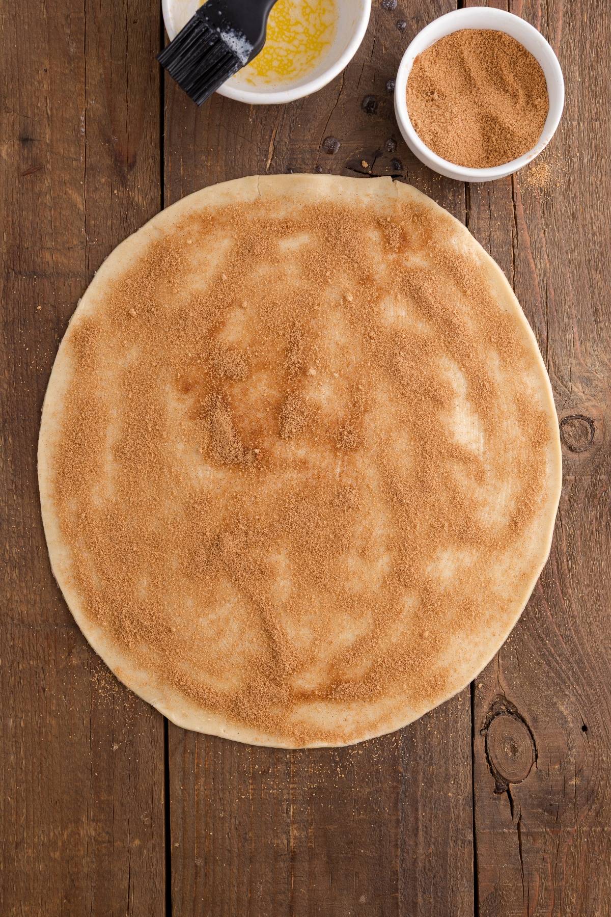 Round pizza dough topped with cinnamon sugar on a wooden surface, with bowls of melted butter and cinnamon sugar nearby.