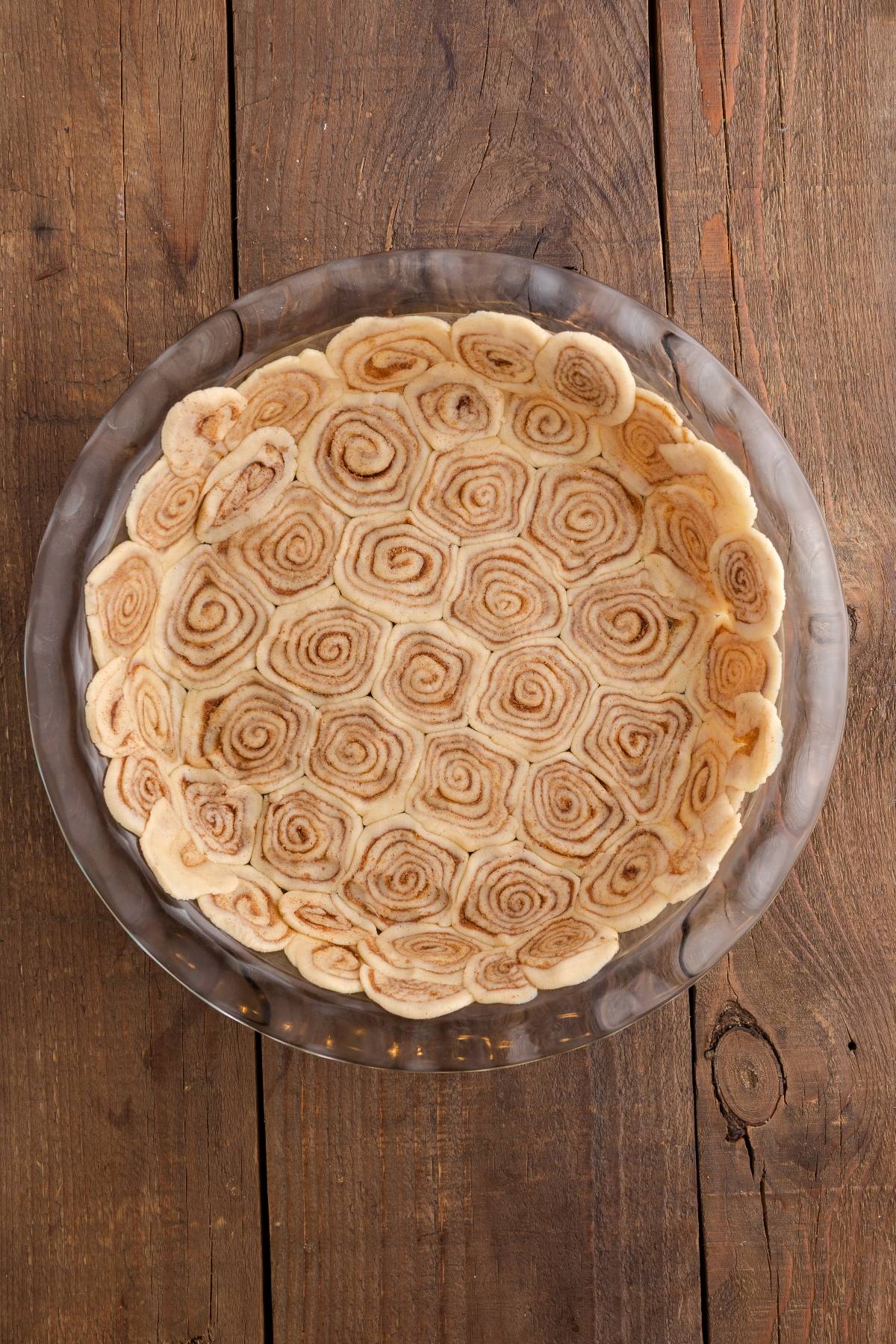 A pie crust made of cinnamon roll spirals in a glass pie dish on a wooden surface.