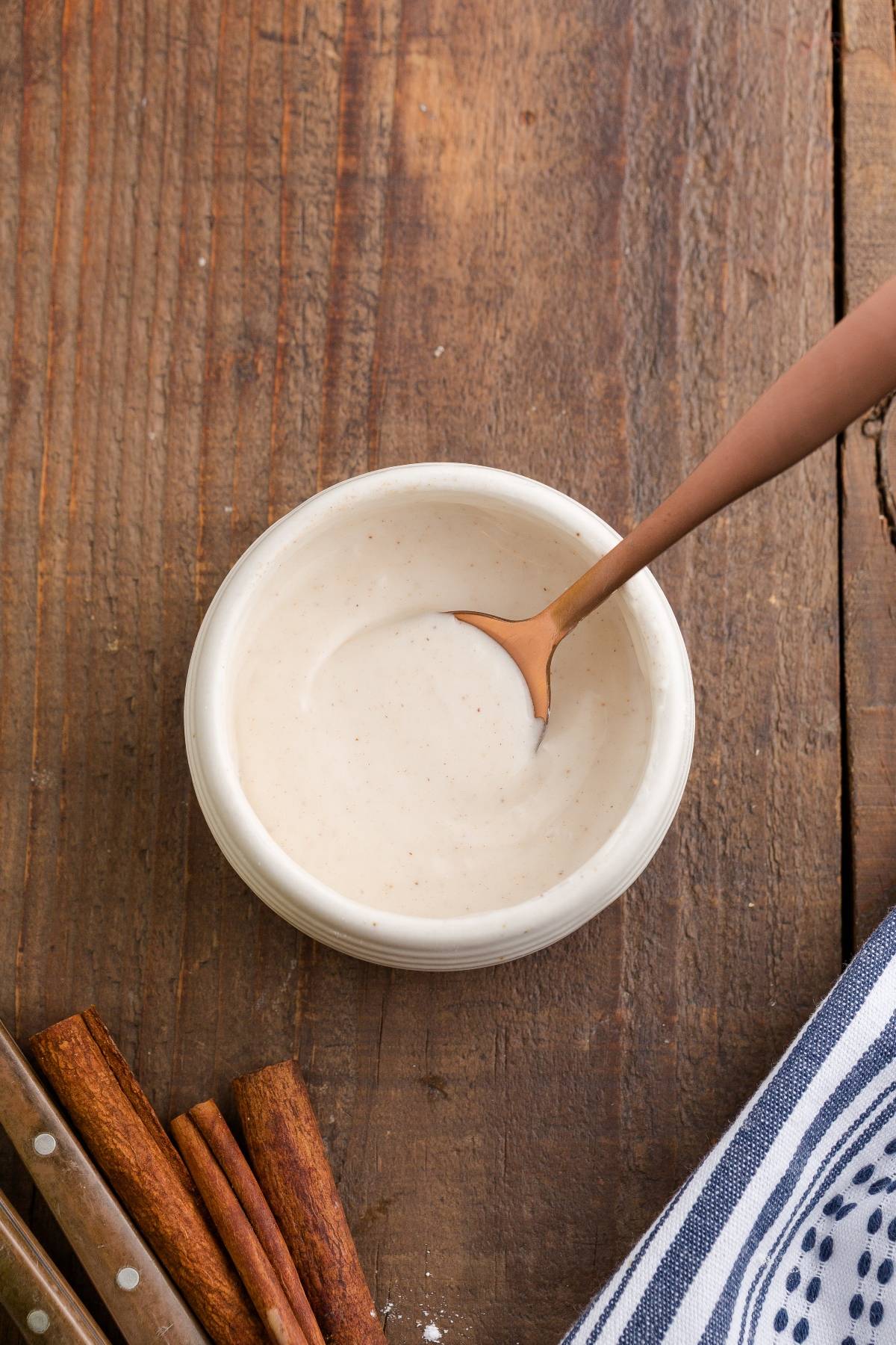 A small bowl of creamy sauce with a spoon, next to cinnamon sticks on a rustic wooden table.