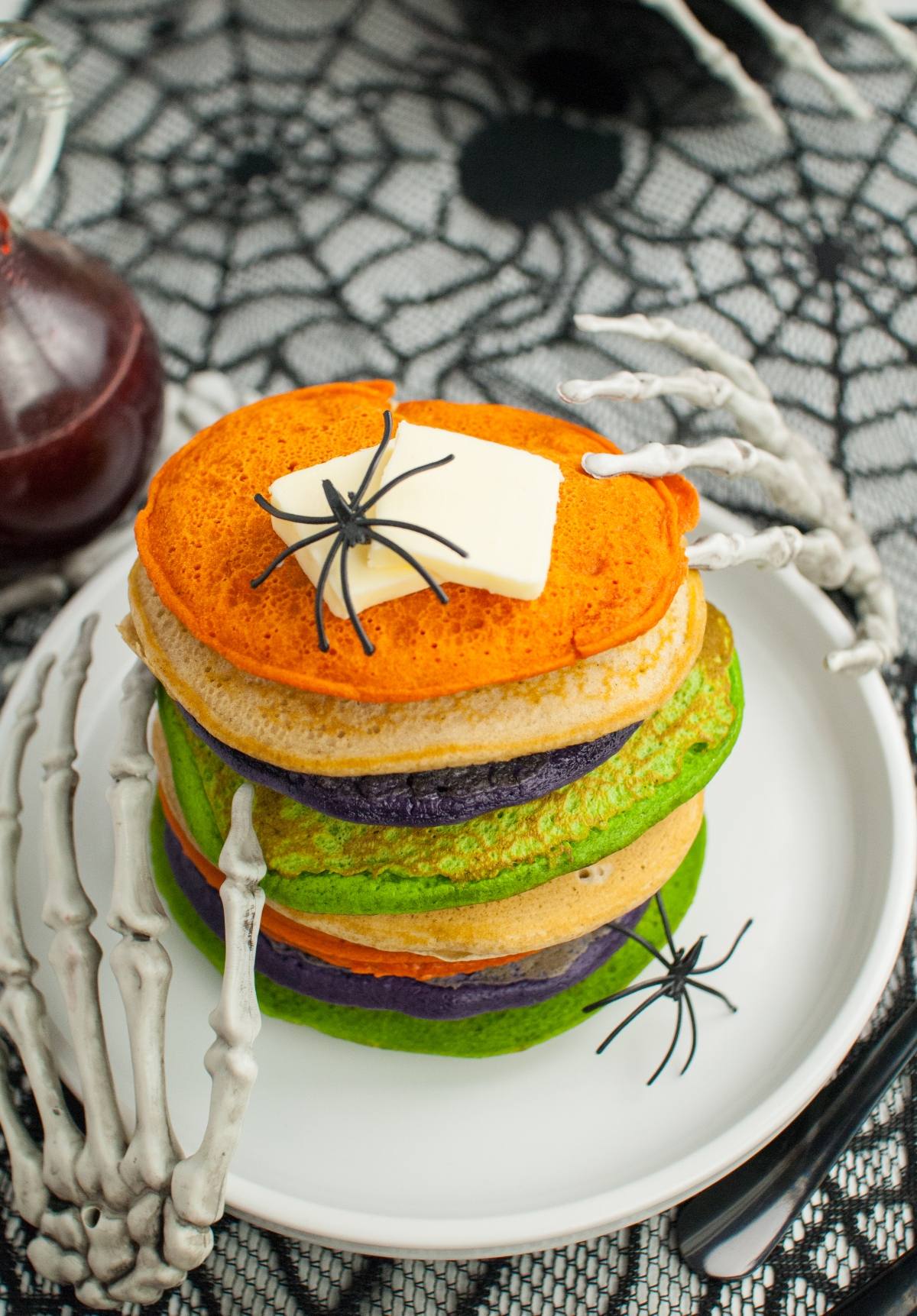 Stack of colorful Halloween pancakes topped with butter, fake spiders, and skeleton hands on a spiderweb tablecloth—perfect for spooky breakfast ideas or Halloween party food!.