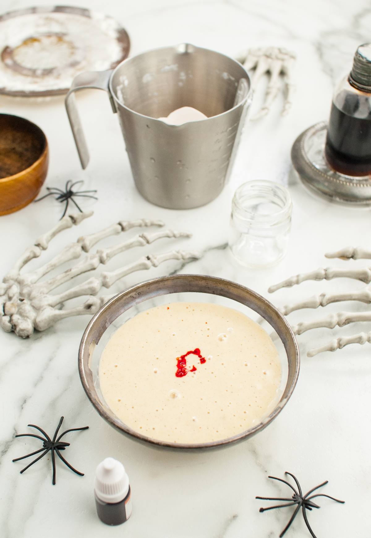 Bowl of batter with red food coloring, surrounded by skeleton hands and Halloween decorations on a marble surface—perfect for making Colorful Halloween Pancakes or trying fun Halloween recipes for kids.