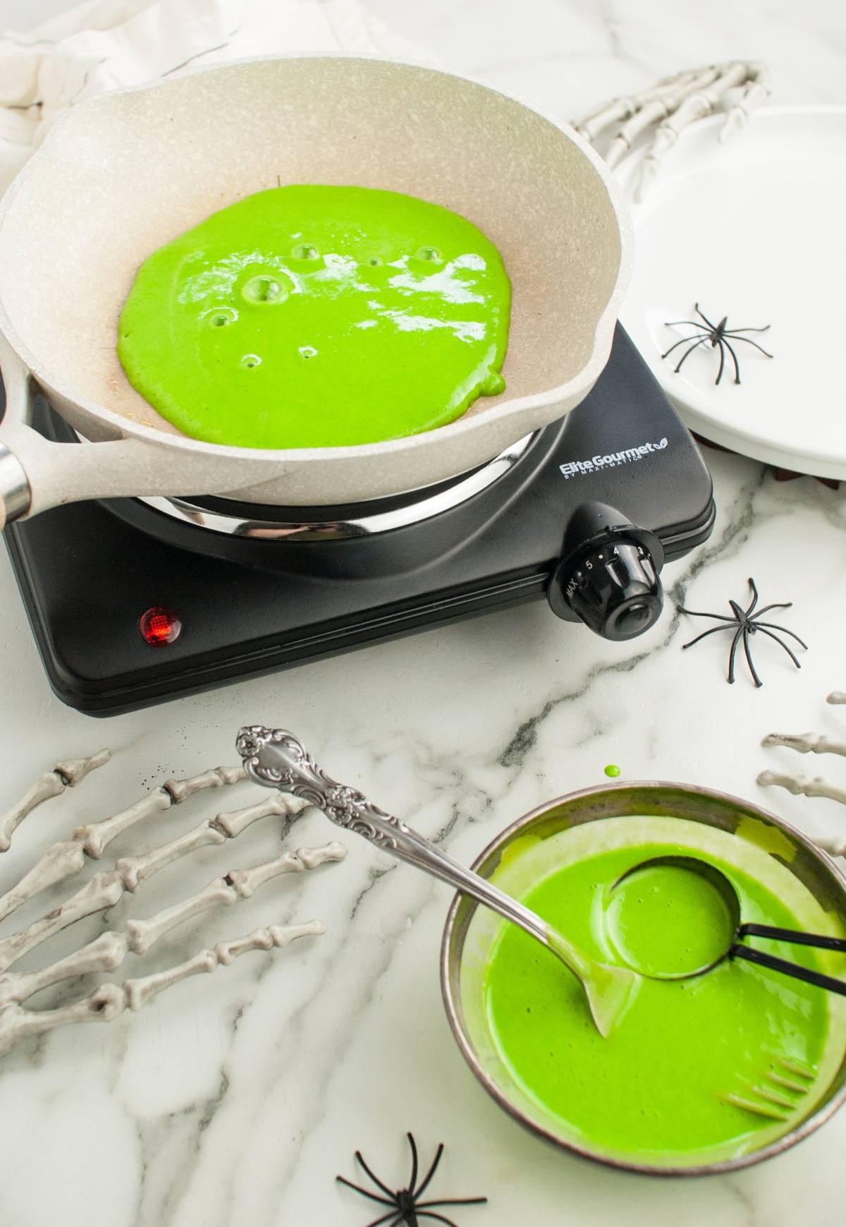 Green pancake batter cooking in a skillet, with a bowl of batter and Halloween decorations nearby—perfect for anyone looking for how to make colourful pancakes or fun Colorful Halloween Pancakes for your next Halloween party food spread.