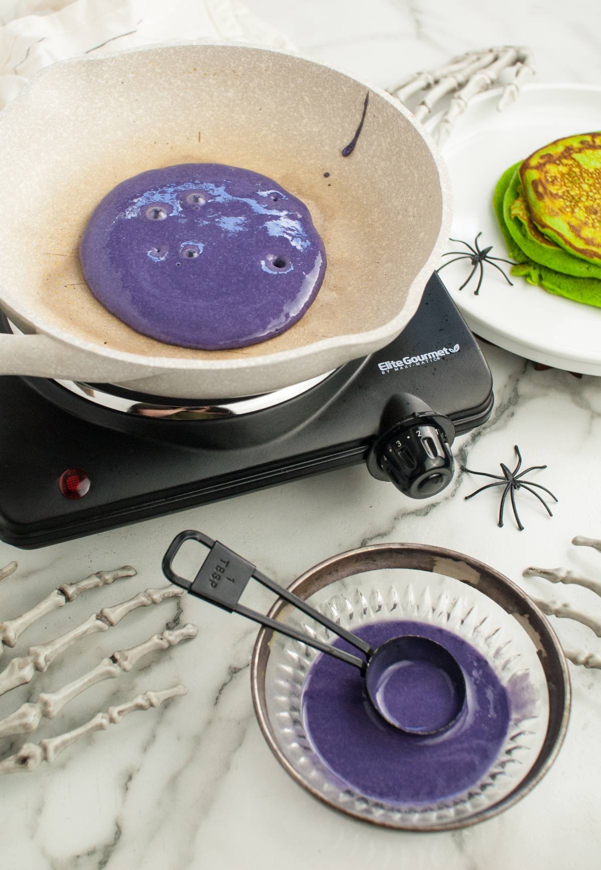 Purple pancake batter cooking in a pan, with green pancakes, spiders, and skeleton hands nearby on a marble surface—perfect for colorful Halloween pancakes and spooky Halloween recipes for kids.
