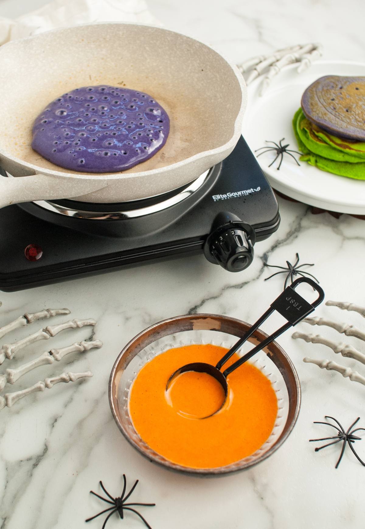 Purple pancake cooking in a pan, with green pancakes and orange batter nearby on a marble surface with spider decor—perfect for your Halloween party food spread or as fun, colorful Halloween pancakes kids will love.