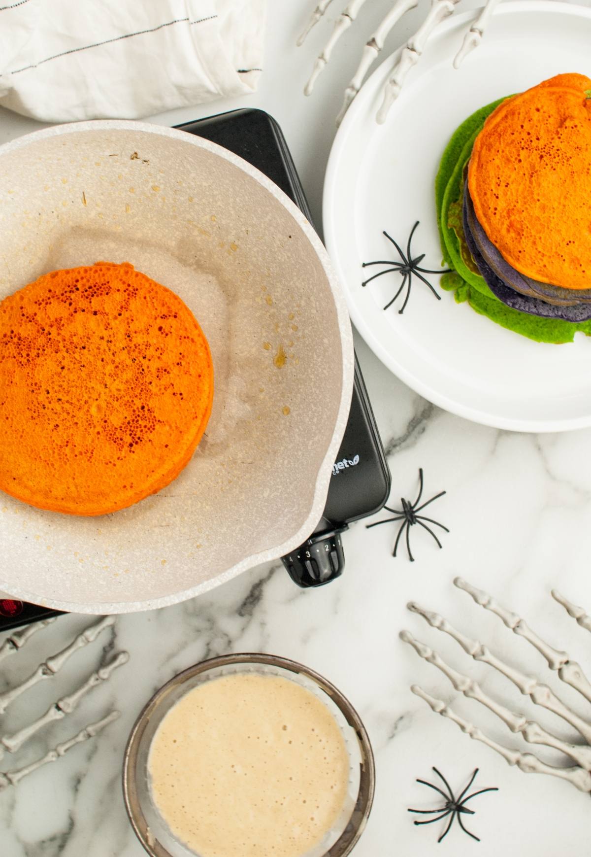 Colorful Halloween Pancakes cooking in a pan, with a vibrant stack of orange, green, and purple pancakes on a plate nearby—perfect Halloween party food for learning how to make colourful pancakes at home.