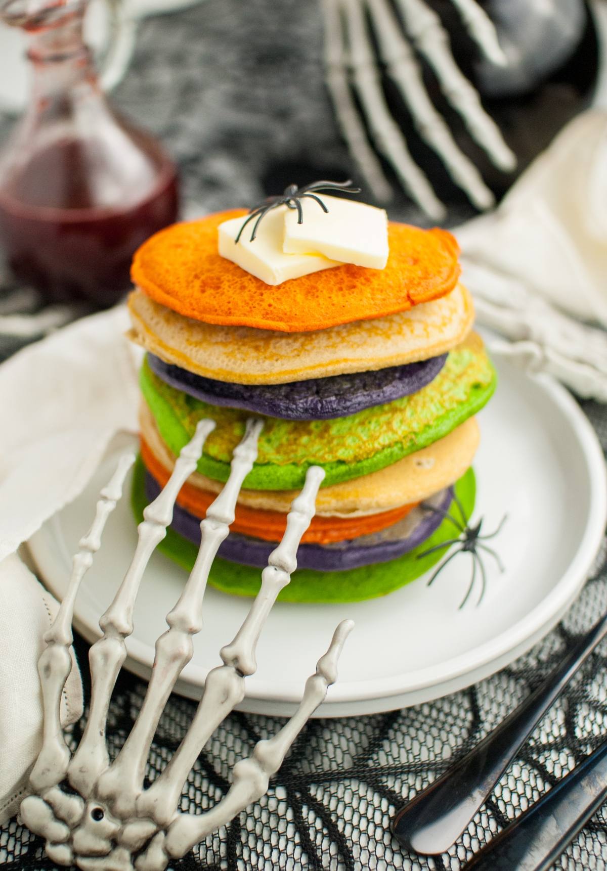 A skeleton hand holds Colorful Halloween Pancakes topped with butter and a fake spider on a white plate—spooky Halloween party food that's as fun as it is tasty!.