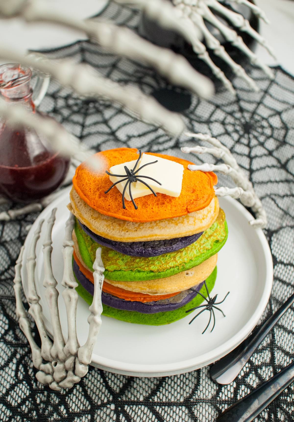 Colorful pancakes topped with butter and toy spiders, held by skeleton hands on a Halloween-themed table—spooky breakfast ideas perfect for Halloween recipes for kids.