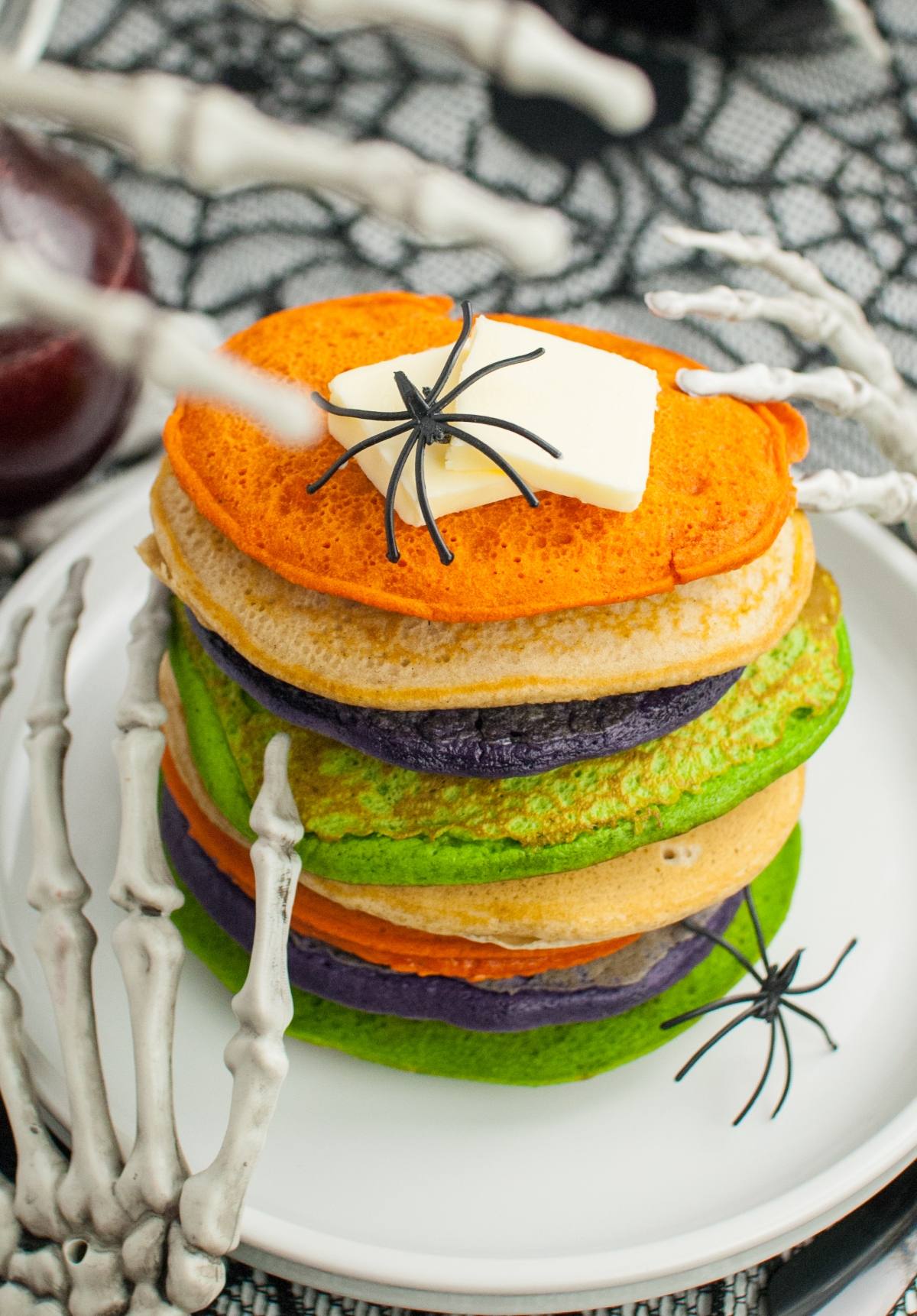 Stack of colorful pancakes with fake spiders, butter on top, and skeleton hands holding the plate—perfect Halloween party food for kids who love spooky treats!.