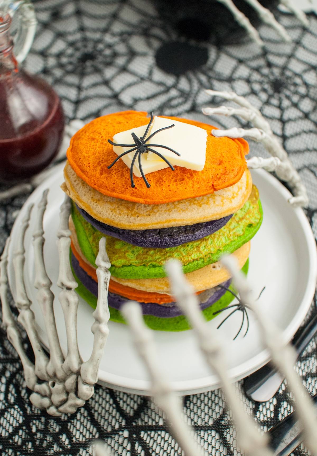 Stack of colorful Halloween pancakes, topped with butter and a fake spider, held by skeleton hands—perfect for spooky breakfast ideas or as fun Halloween party food.