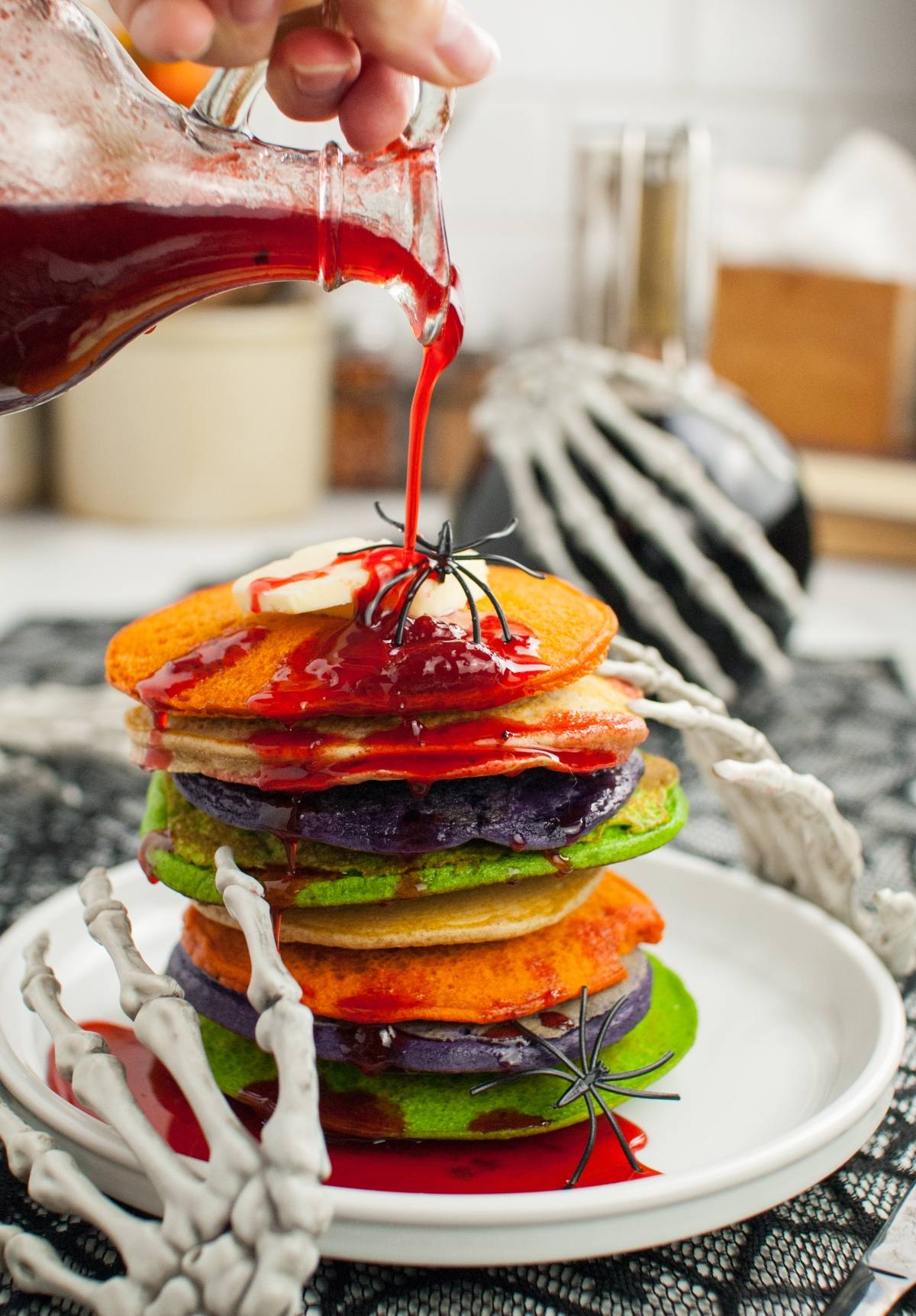 A stack of colorful pancakes with syrup being poured, topped with fake spiders and skeleton hands—perfect Halloween party food and a fun idea for Halloween recipes for kids.