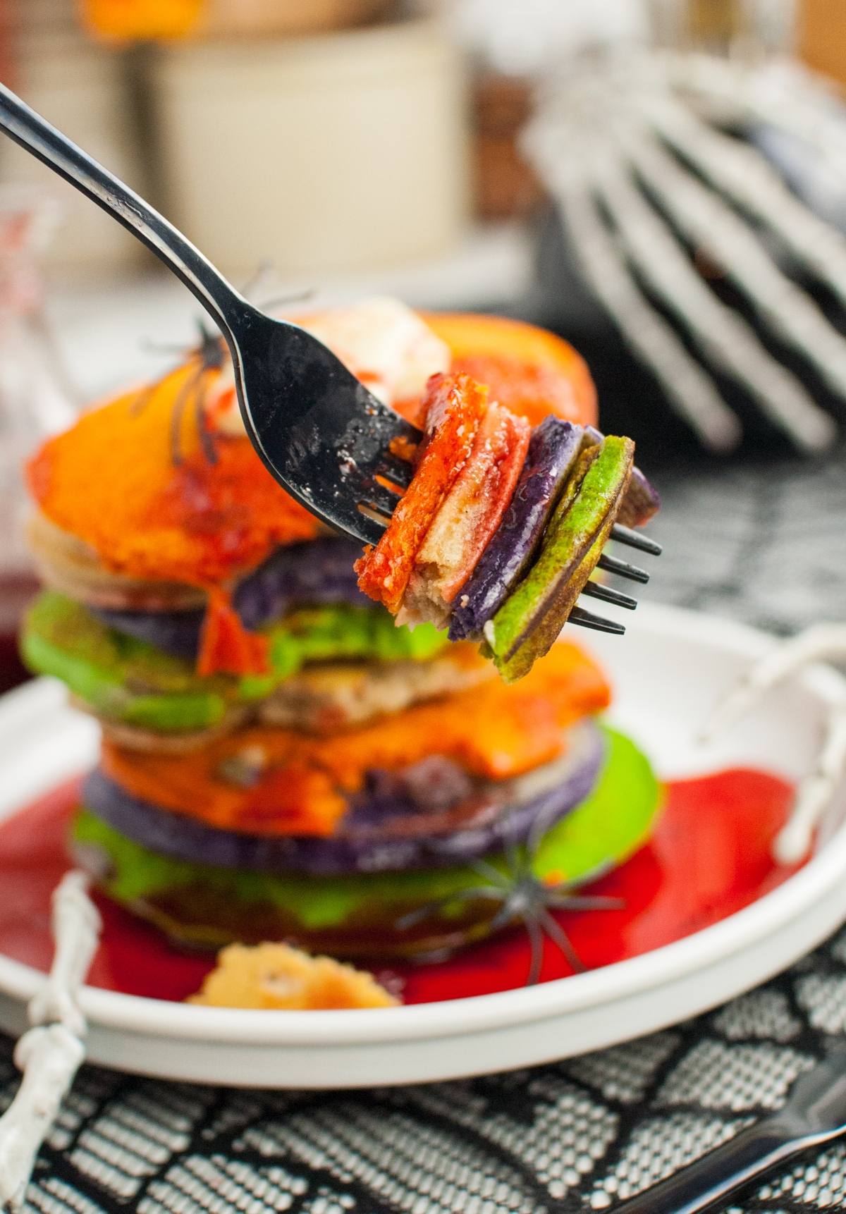 A fork holds a bite of Colorful Halloween Pancakes; the vibrant stack sits on a plate with red syrup and Halloween decor—perfect for spooky breakfast ideas.