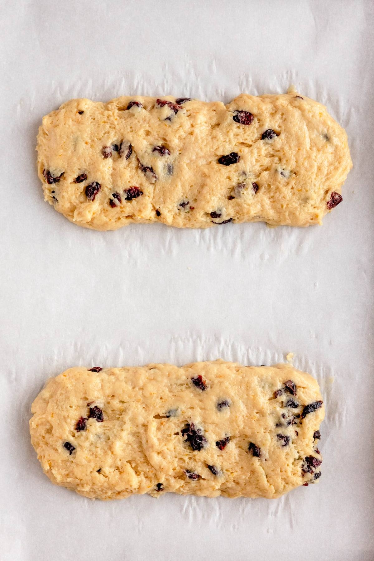 Two unbaked cranberry orange biscotti dough logs with dried fruit rest on parchment paper, ready for baking.