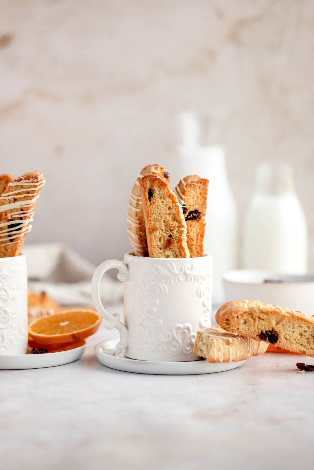 White mugs filled with cranberry orange biscotti on saucers, with milk bottles and orange halves in the background.