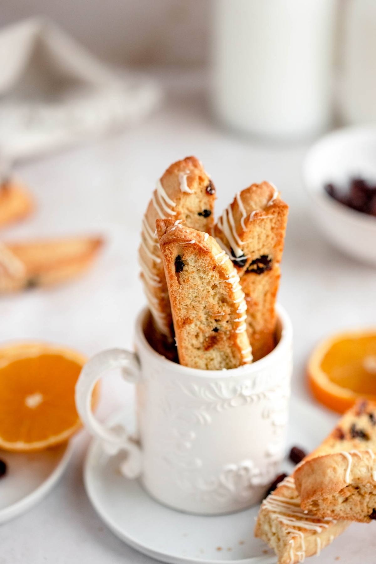 Three cranberry orange biscotti with white drizzle stand upright in a decorative white mug, surrounded by fresh orange slices.