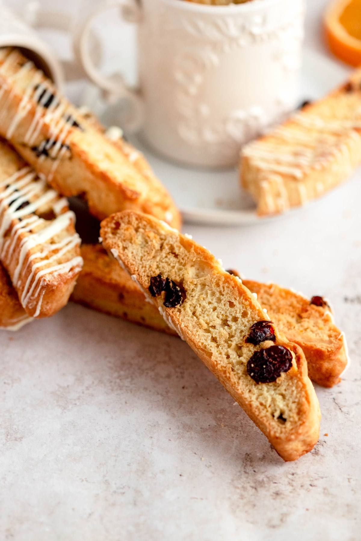 Cranberry orange biscotti stacked on a table with white drizzle and a cup in the background.