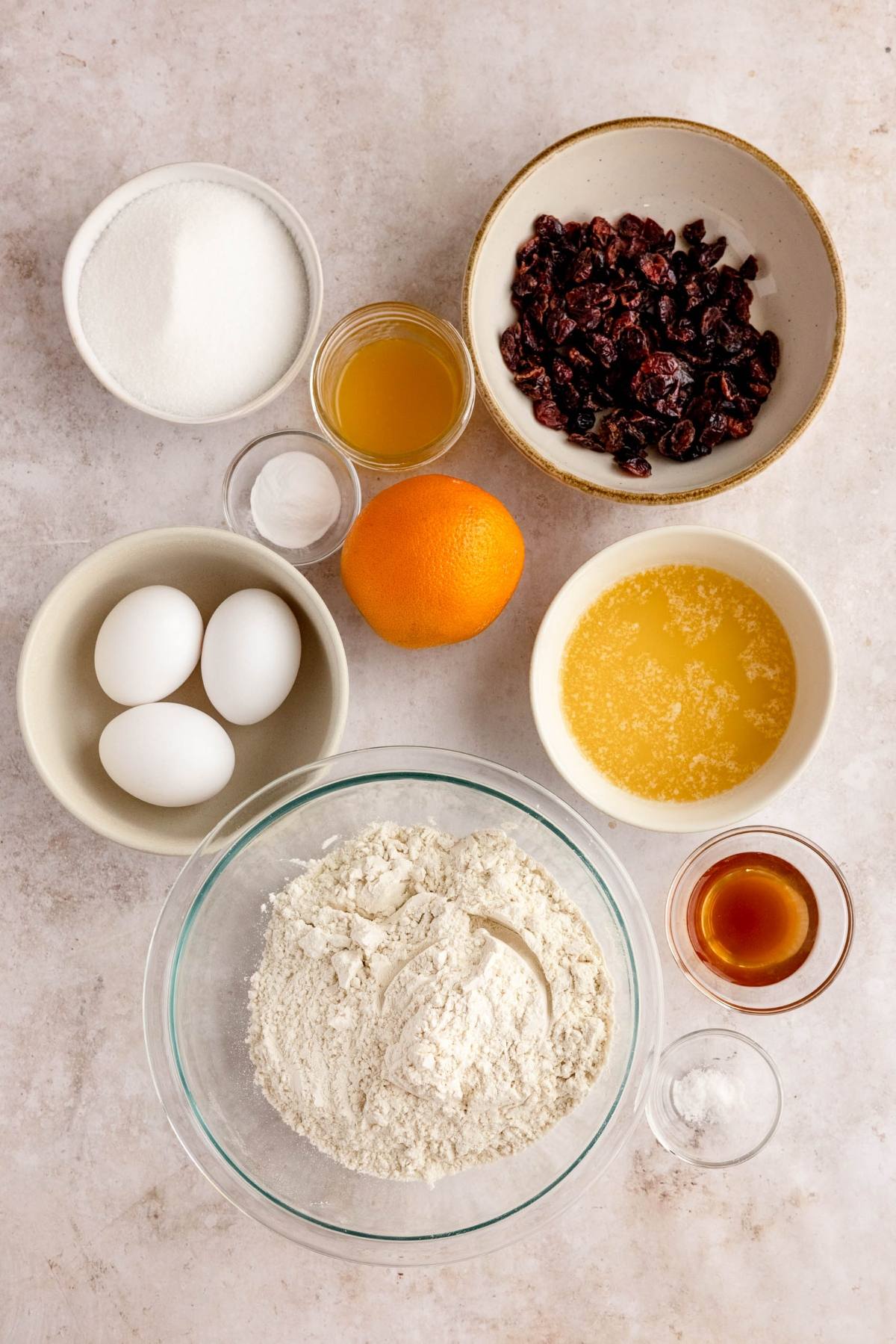 Bowls of flour, sugar, dried cranberries, eggs, orange, and other baking essentials on a light surface—perfect for making cranberry orange biscotti.