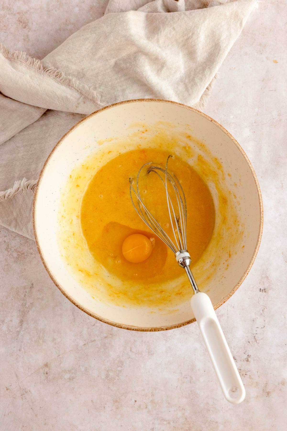 A mixing bowl with a whisk, beaten eggs, and one unbroken egg yolk sits on a light countertop with a beige cloth—just the right start for making cranberry orange biscotti.
