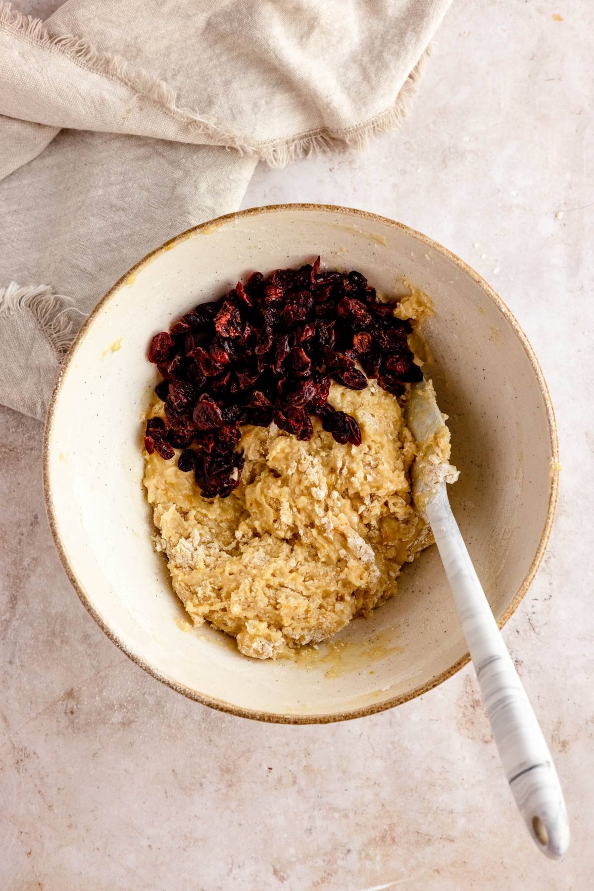 A mixing bowl with cookie dough for cranberry orange biscotti, studded with dried cranberries, and a spatula resting inside.