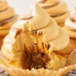 A dulce de leche cupcake with caramel filling and swirled frosting, partially eaten, sits on a wire rack.