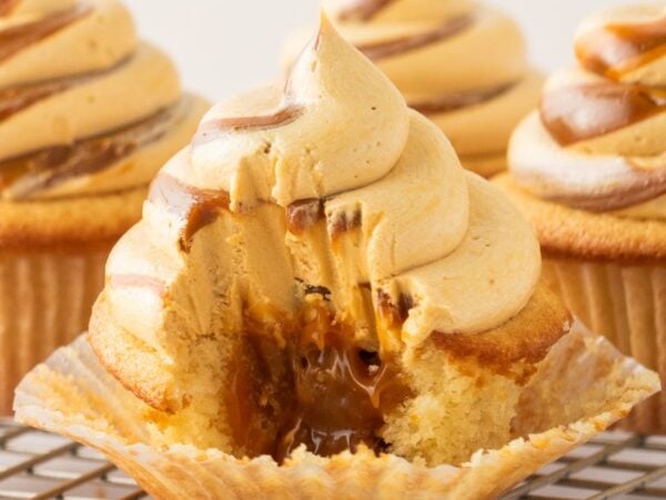A dulce de leche cupcake with caramel filling and swirled frosting, partially eaten, sits on a wire rack.