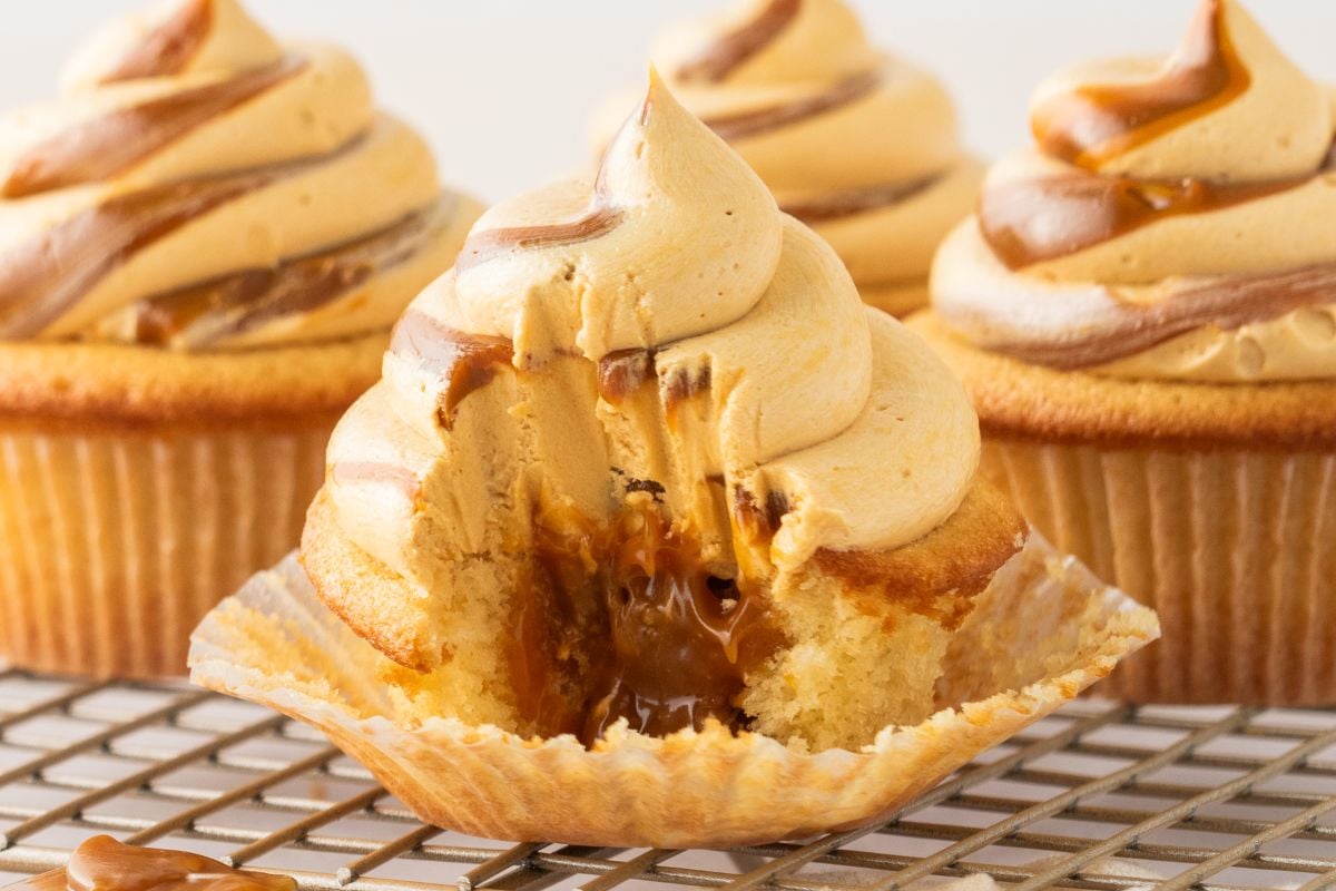 A dulce de leche cupcake with caramel frosting and a gooey caramel center, one bite taken out, sits on a cooling rack.