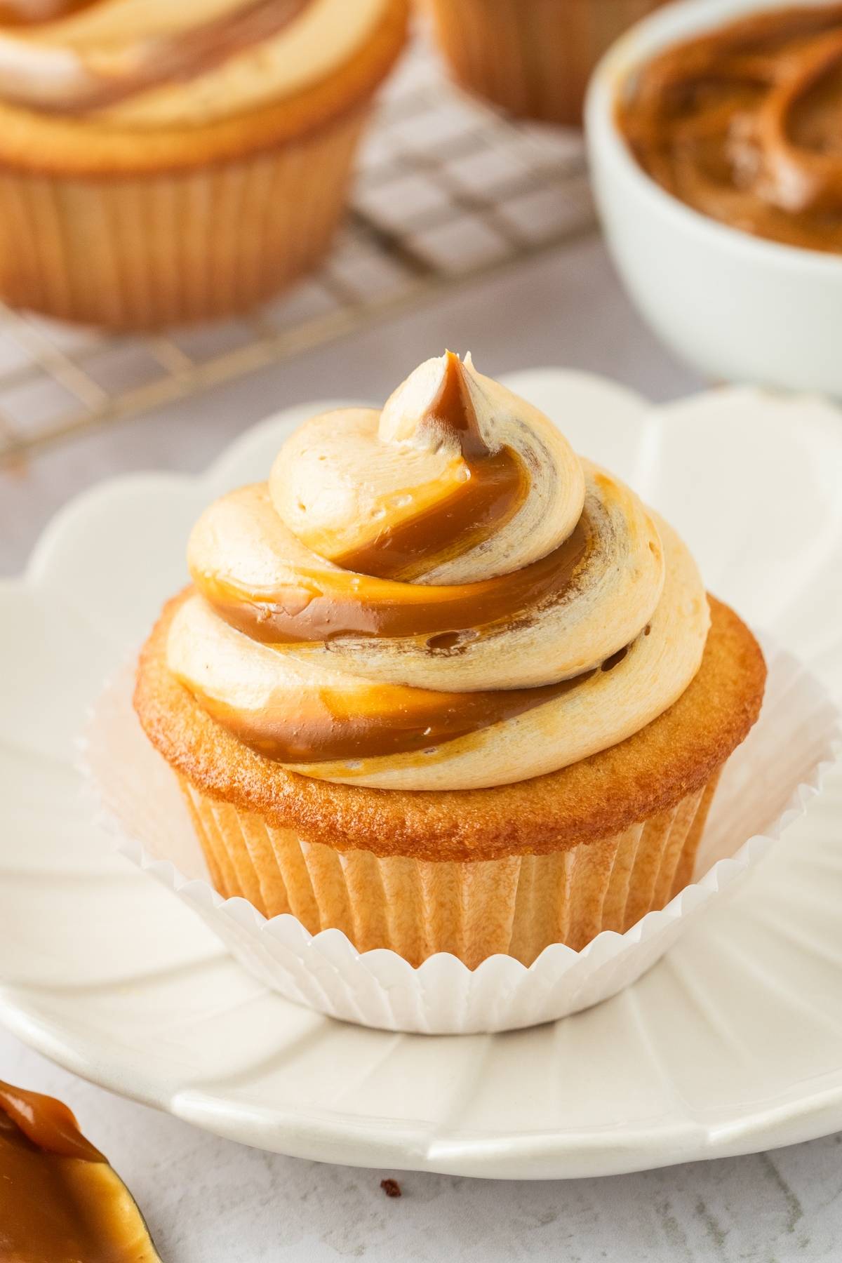 Cupcake with swirled caramel and cream frosting on a white plate, with more cupcakes in the background.