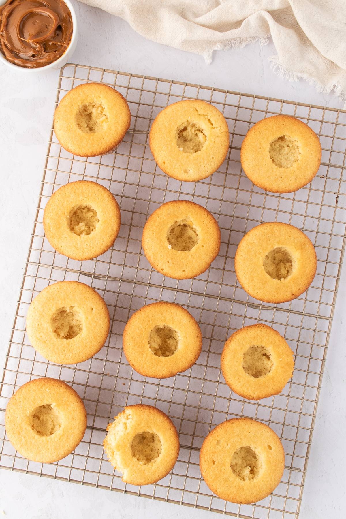 Twelve cupcakes with hollow centers on a cooling rack, next to a small bowl of chocolate spread.