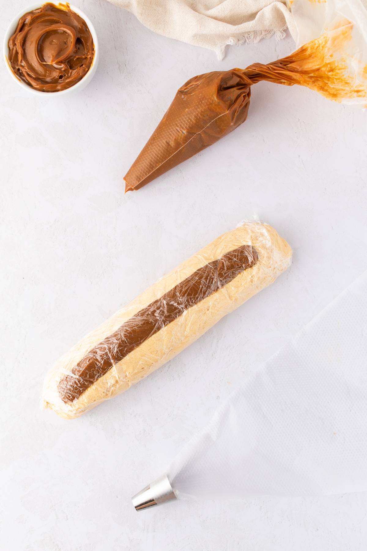 Dough log with a chocolate filling wrapped in plastic, piping bags, and a bowl of spread on a white surface.