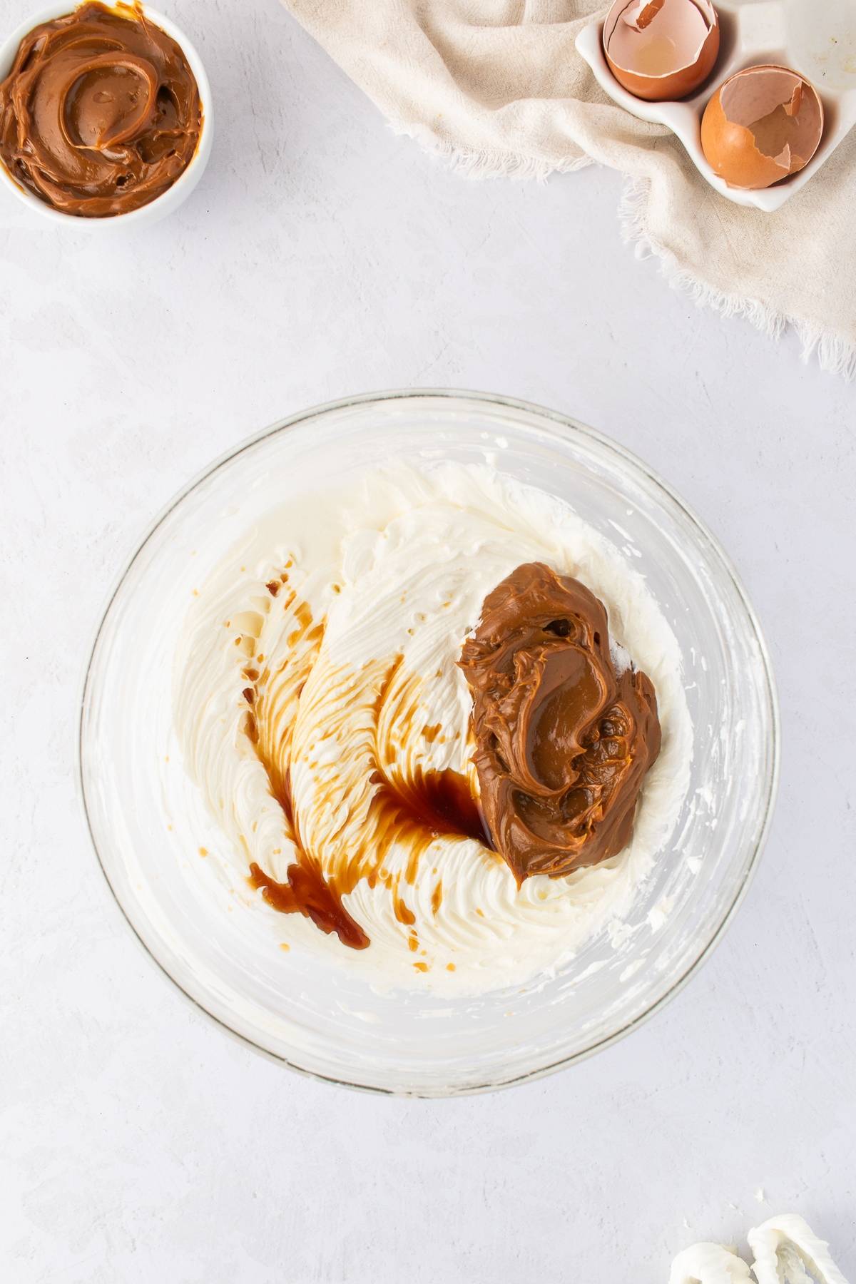 Glass bowl with whipped cream cheese, chocolate spread, and vanilla extract on a white surface with eggs nearby.