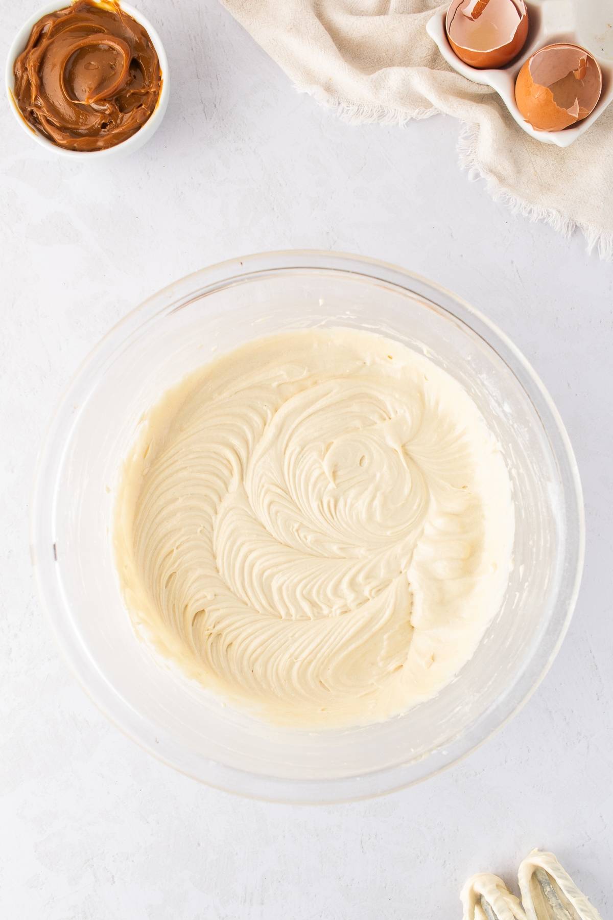 A glass bowl of creamy cake batter next to a bowl of caramel, eggshells, and a kitchen towel.