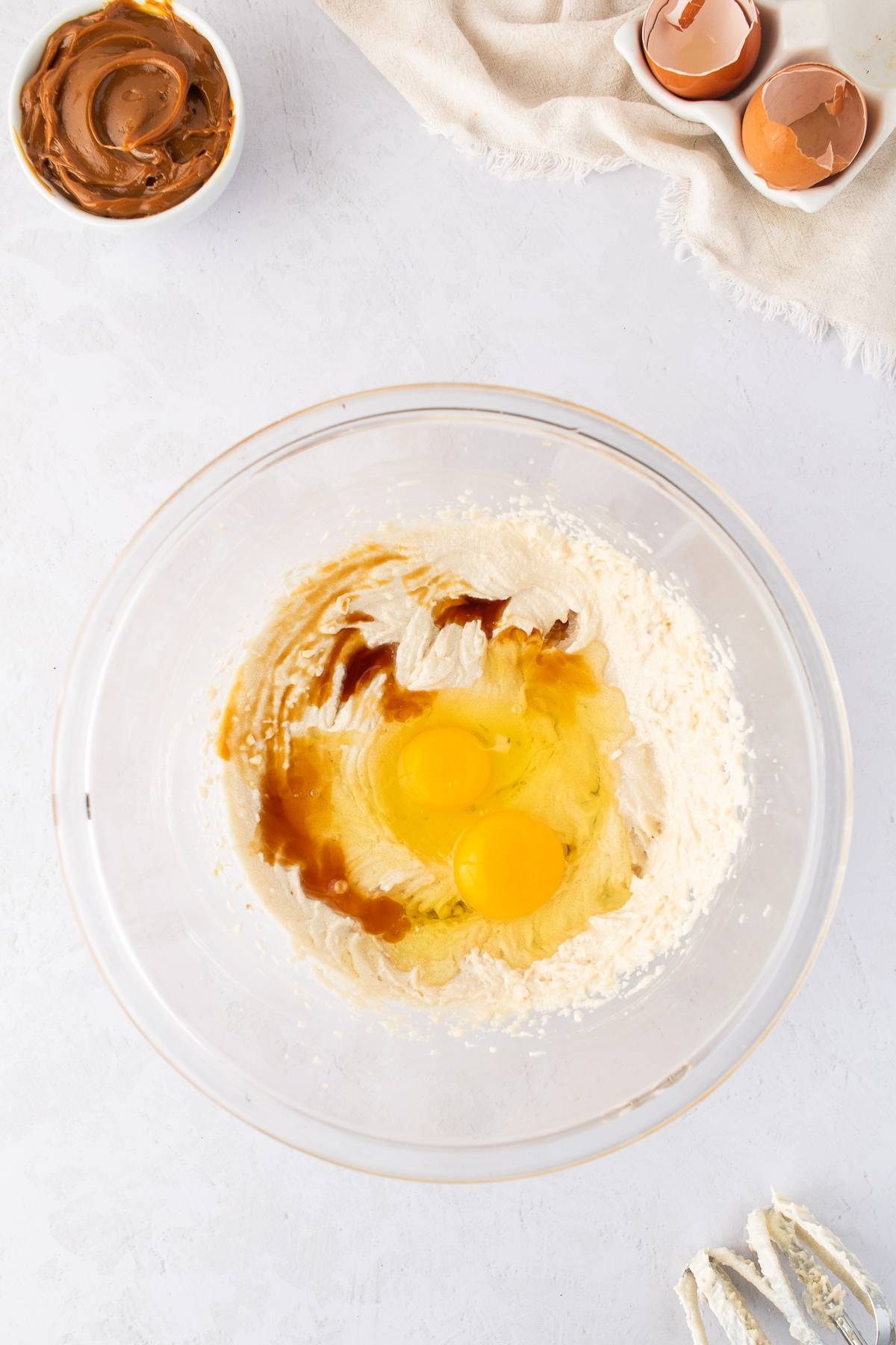 A glass bowl with flour, eggs, and vanilla, next to cracked eggshells and a small bowl of brown spread.