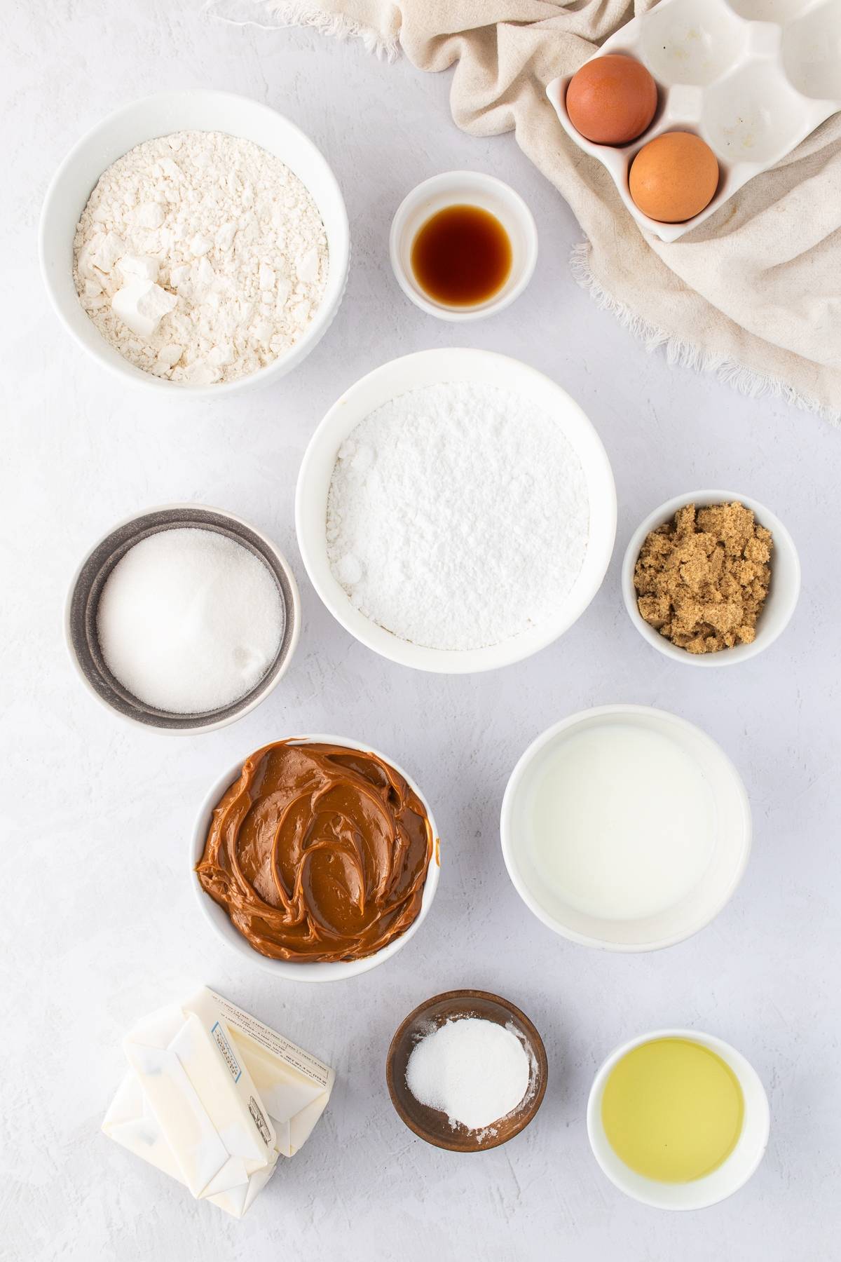 Bowls of flour, sugar, vanilla, brown sugar, eggs, butter, oil, baking powder, and caramel on a white surface.