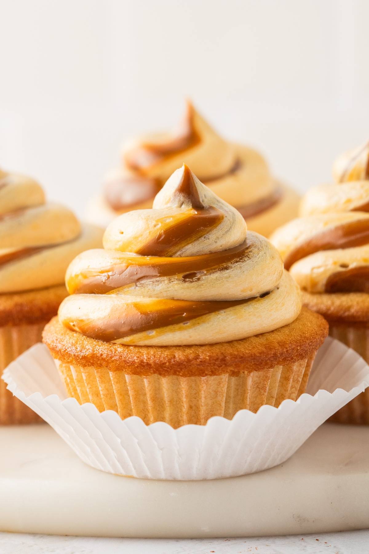 A vanilla cupcake with swirled caramel frosting in a white paper liner, with more cupcakes in the background.