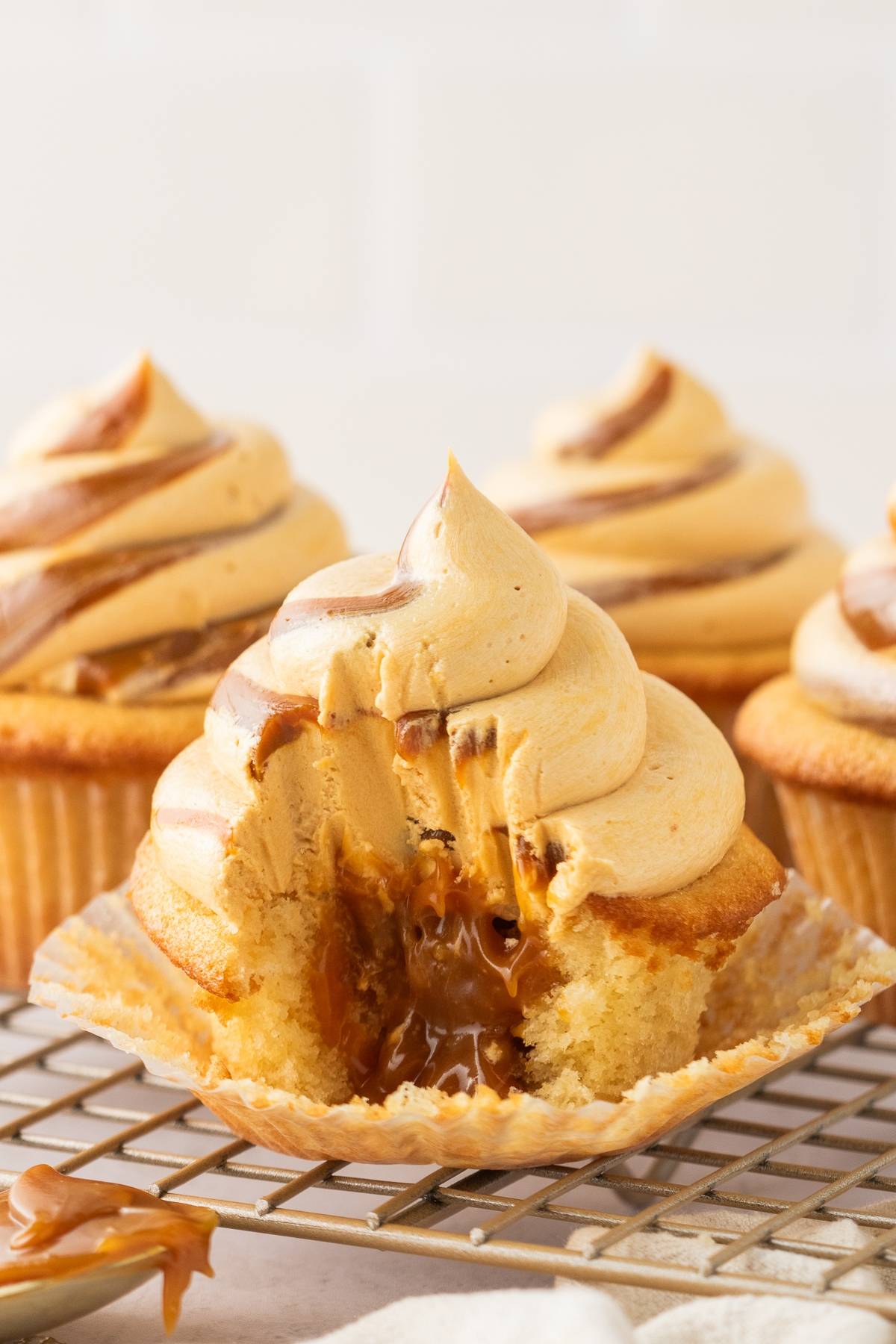 Cupcake with caramel filling and swirled frosting, partially unwrapped and bitten, on a cooling rack.