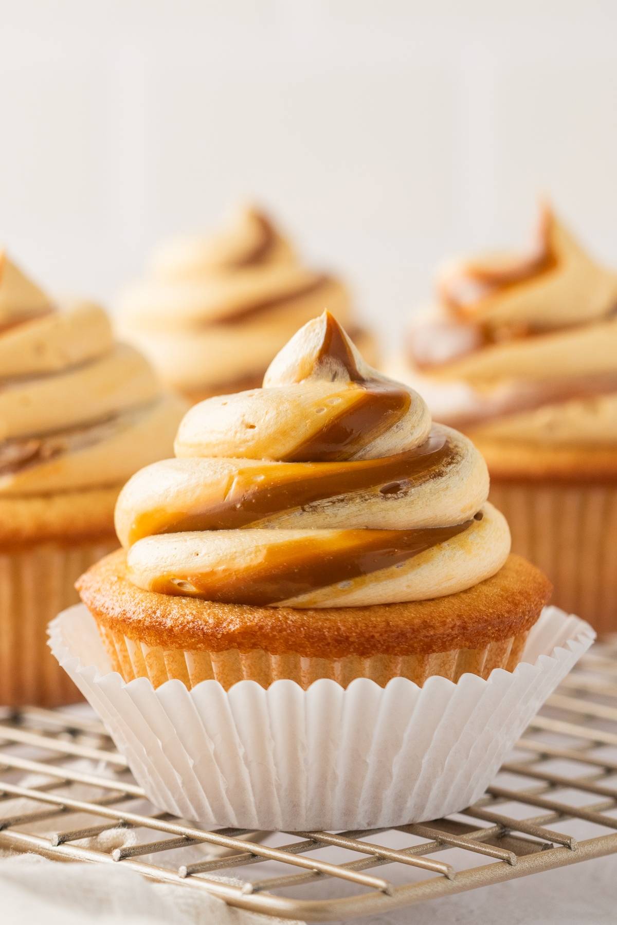 A cupcake with caramel and vanilla swirled frosting on a cooling rack, with more cupcakes in the background.