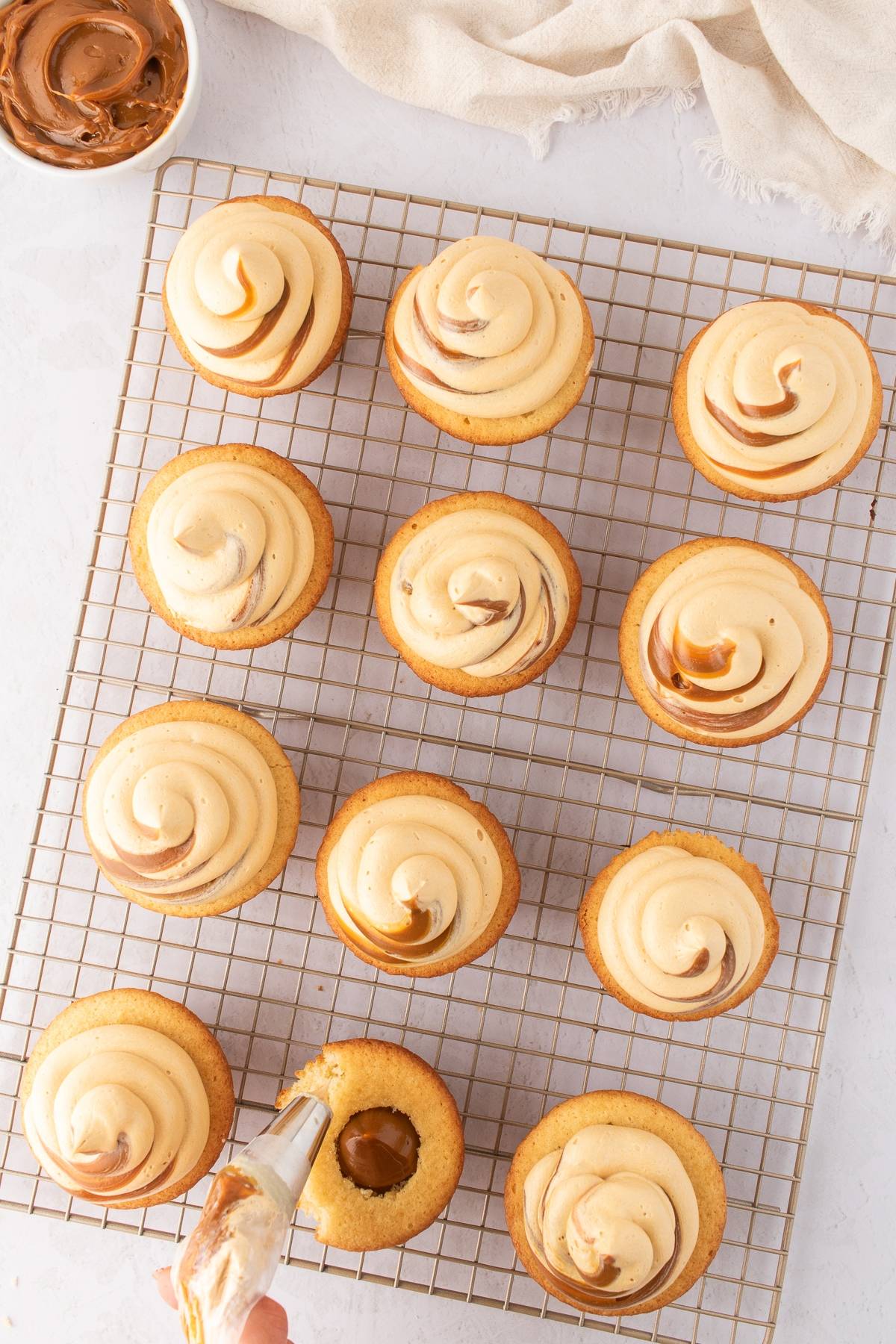 Piping frosting onto cupcakes with swirled icing on a cooling rack; bowl of frosting nearby.