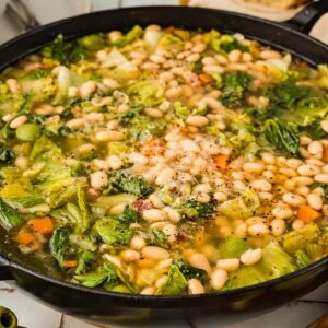 A pot of Escarole and Bean Soup with leafy greens, carrots, white beans, and fresh herbs sits on the table.