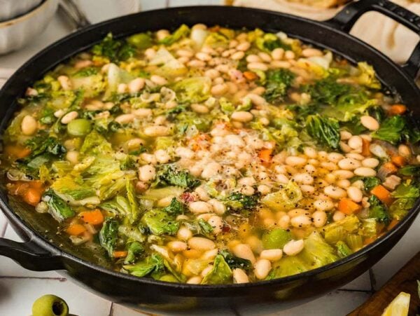 A pot of Escarole and Bean Soup with leafy greens, carrots, white beans, and fresh herbs sits on the table.