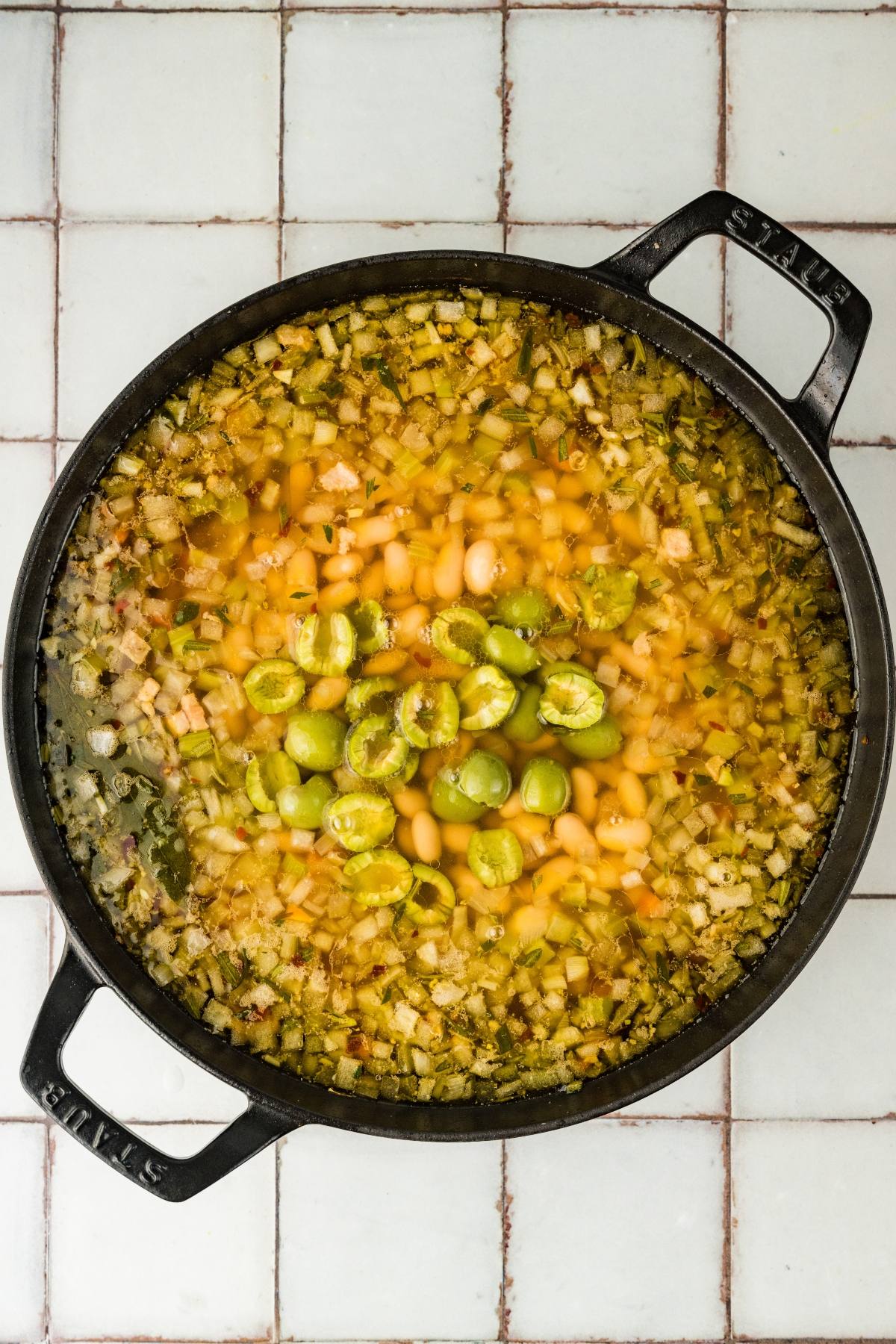 A pot of soup with diced vegetables, white beans, and green olives on a tiled surface.