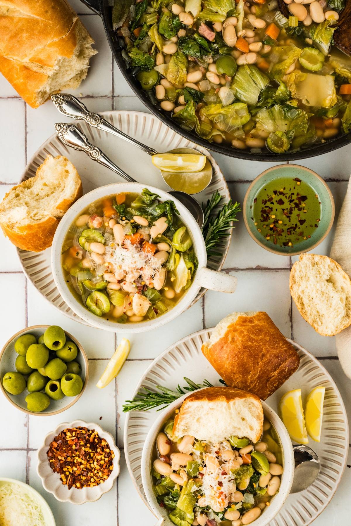 Two bowls of white bean and greens soup with bread, lemon wedges, olives, and seasonings on a tiled table.