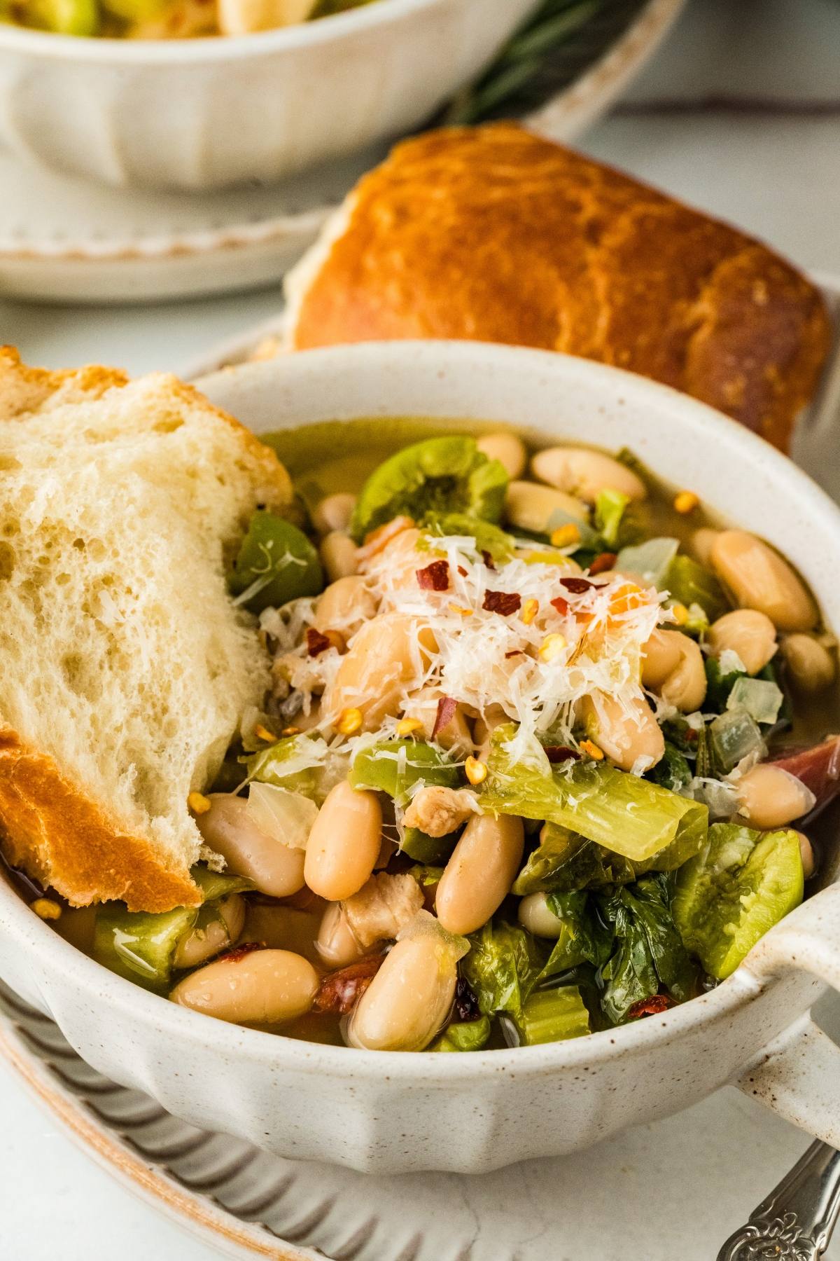 A bowl of white bean and vegetable soup topped with grated cheese, served with a piece of crusty bread.