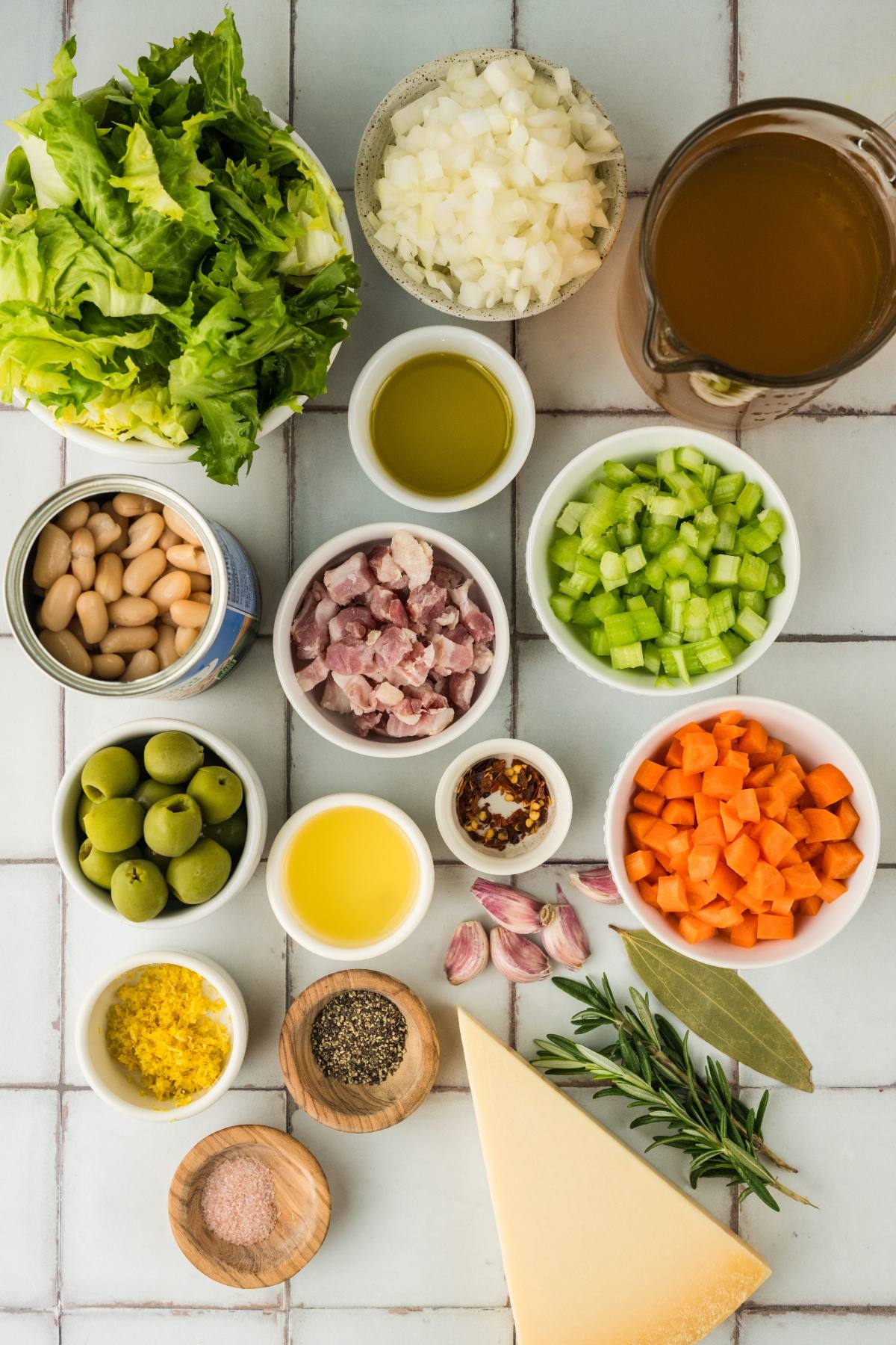Various chopped vegetables, beans, olives, broth, herbs, and seasonings arranged in bowls on a tiled surface.