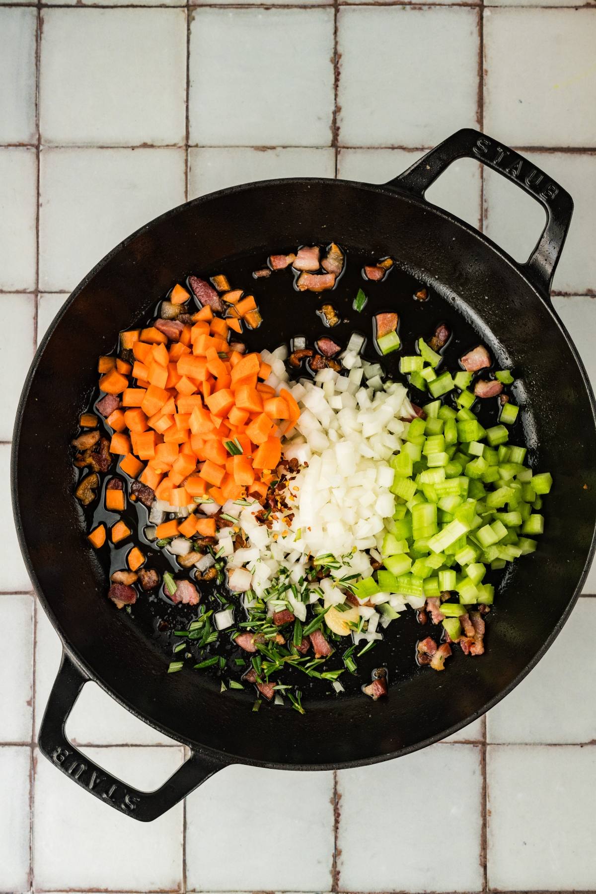 Diced carrots, onions, and celery cooking in a black cast iron pan on a tiled surface.