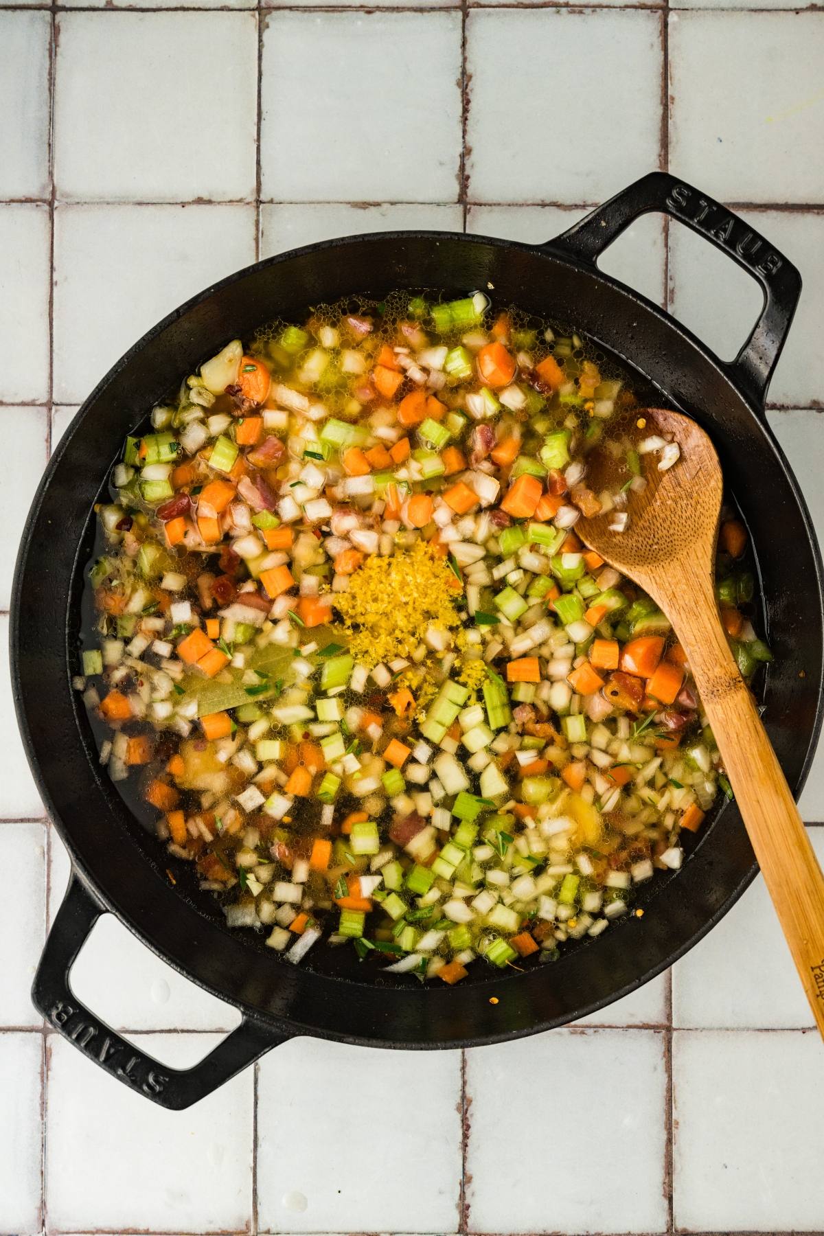 Chopped vegetables and zest simmering in a black pot with a wooden spoon on a tiled surface.