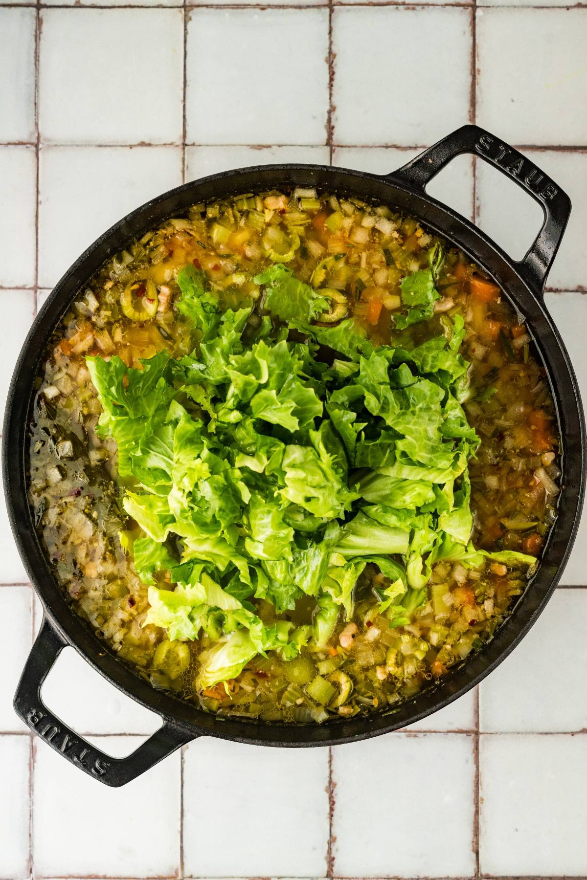 A pot of soup with chopped vegetables and leafy greens on top, sitting on a tiled surface.