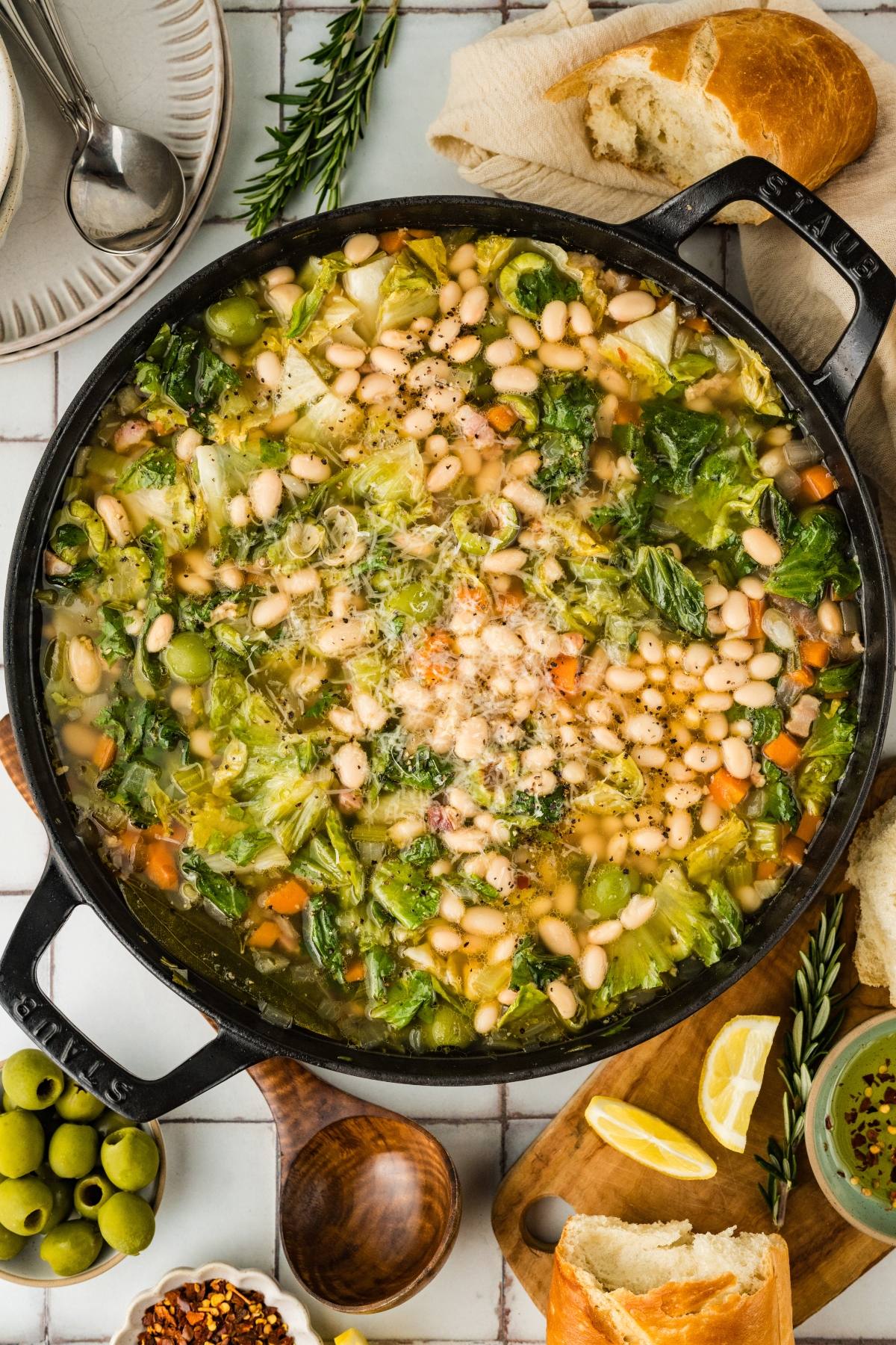 A pot of white bean and vegetable soup with greens, surrounded by bread, olives, lemon, and herbs.