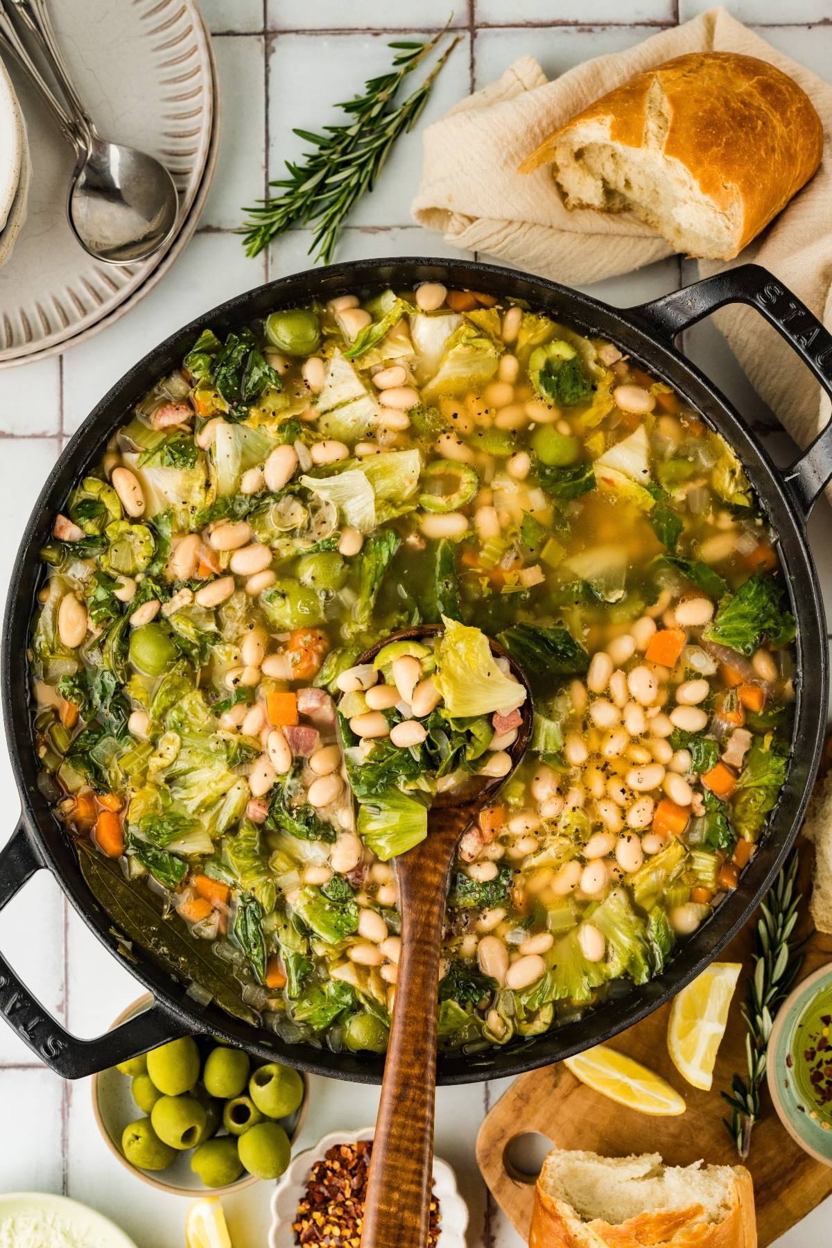 A pot of vegetable and white bean soup with greens, surrounded by bread, olives, and lemon wedges.