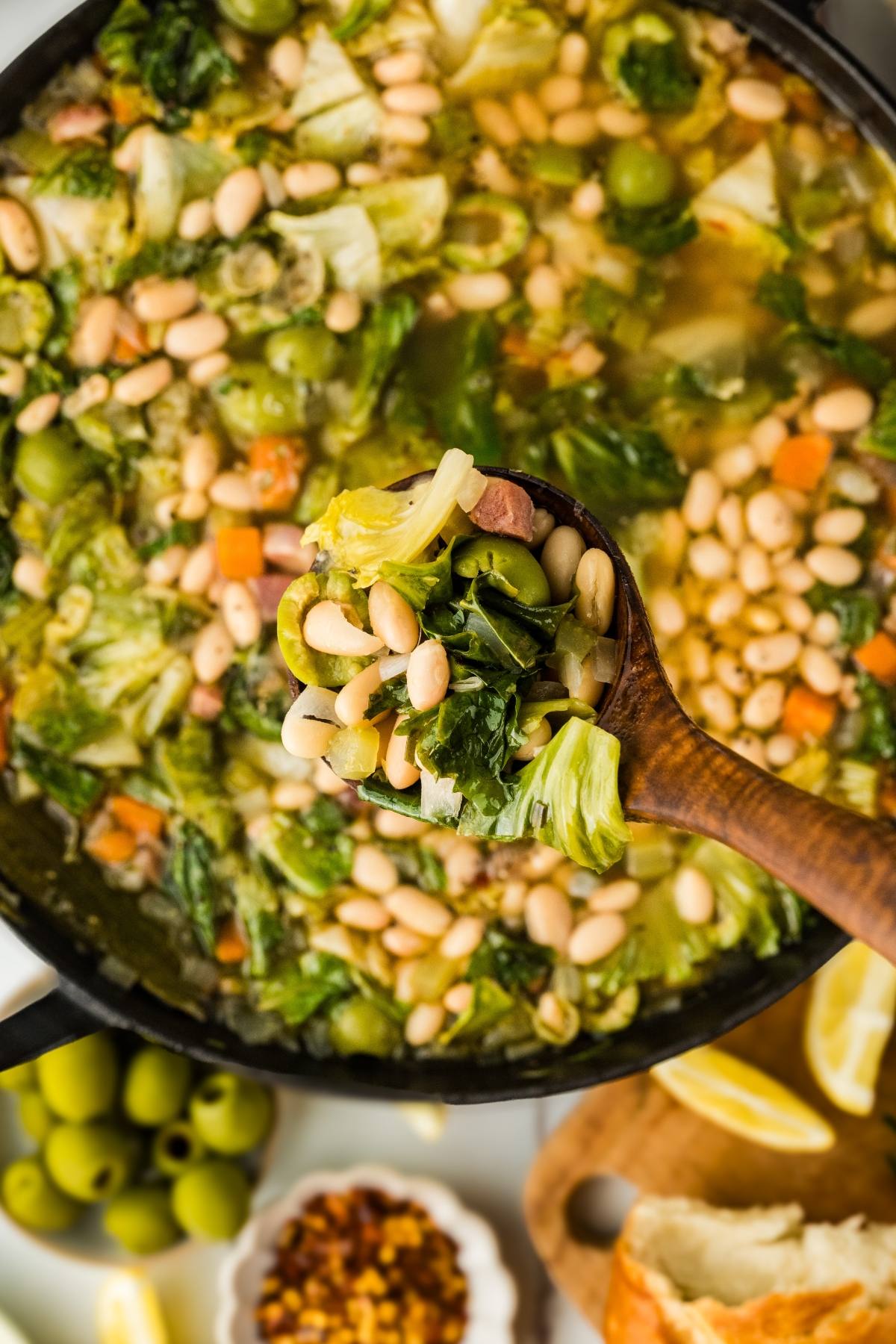 A wooden spoon holds white bean and vegetable soup over a pot, with olives and bread on the side.